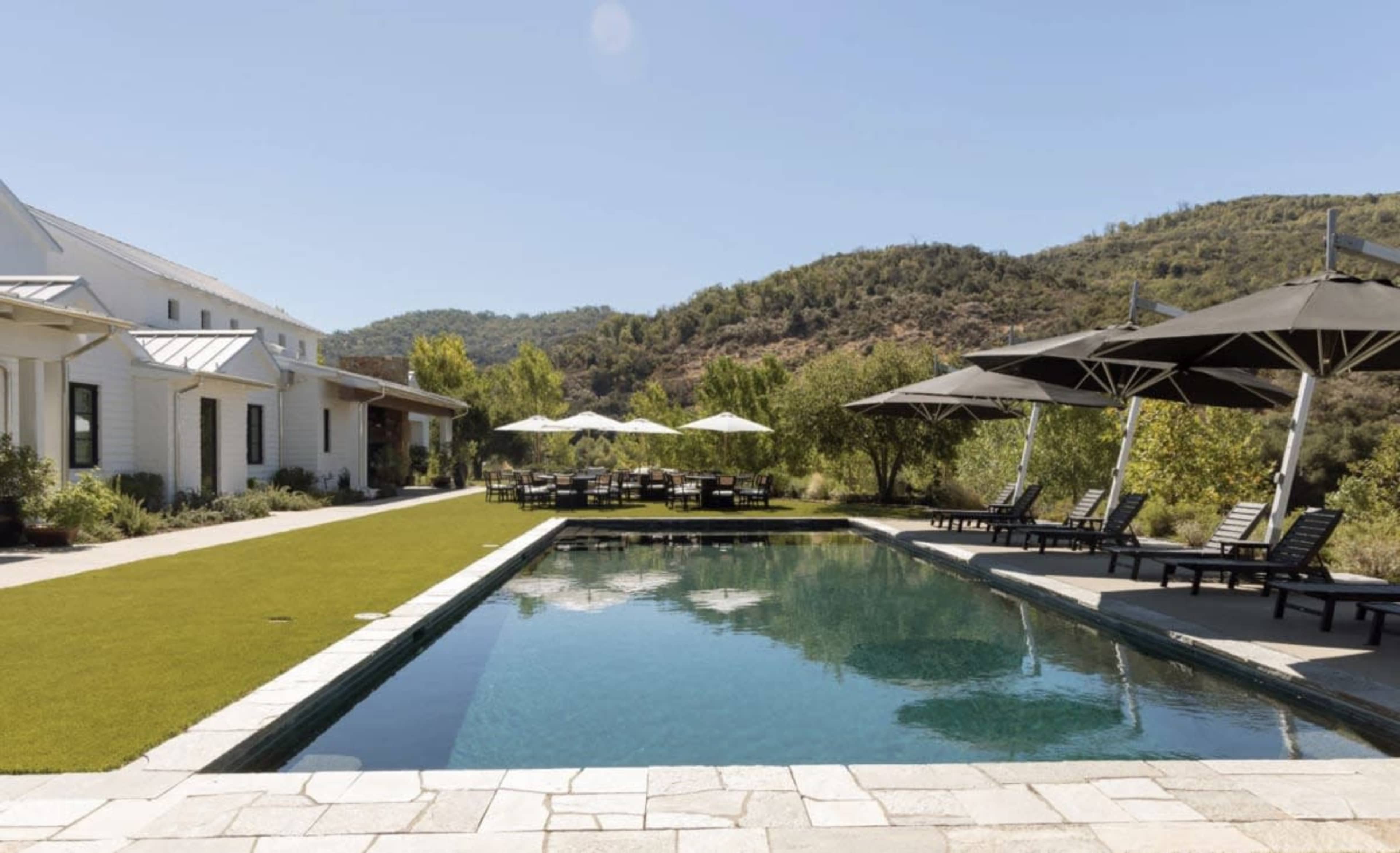 The image shows a modern outdoor space featuring a swimming pool surrounded by lounge chairs, picnic tables, and green landscaping against a backdrop of rolling hills.