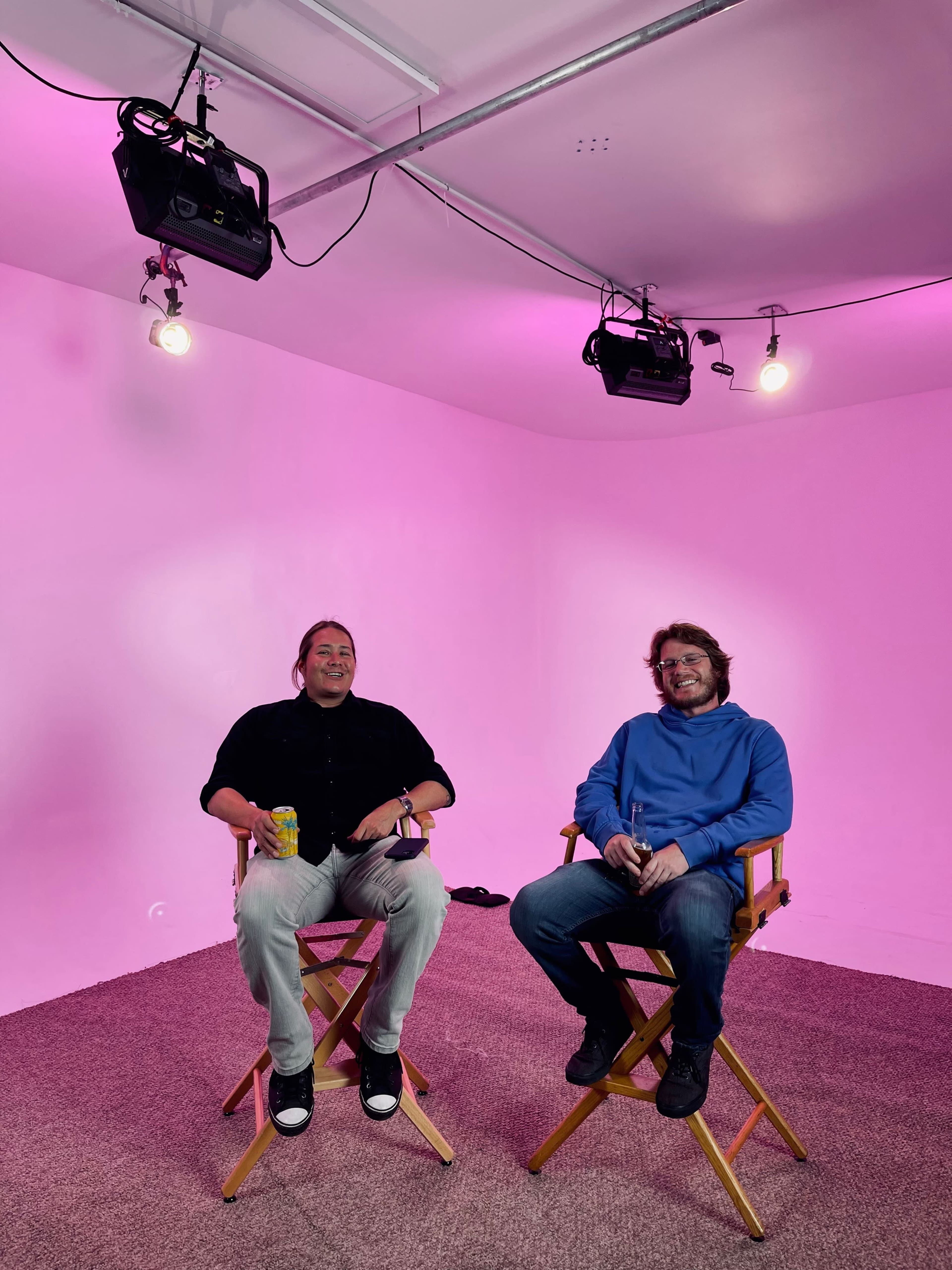 Two men sit on director's chairs in a brightly lit room with pink walls and overhead lights.