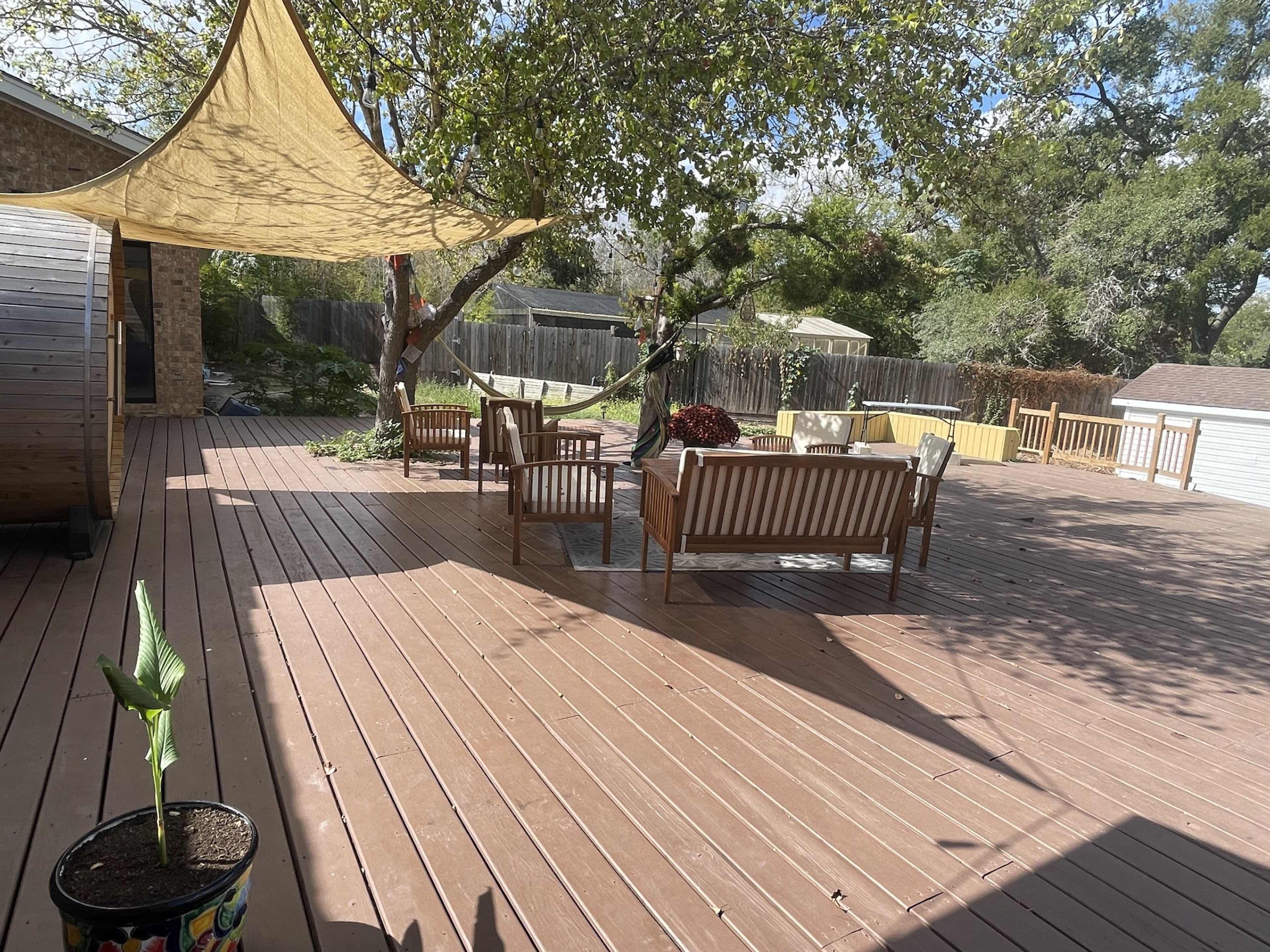 A wooden deck features several seating arrangements and a shade sail above, surrounded by trees and a storage shed.