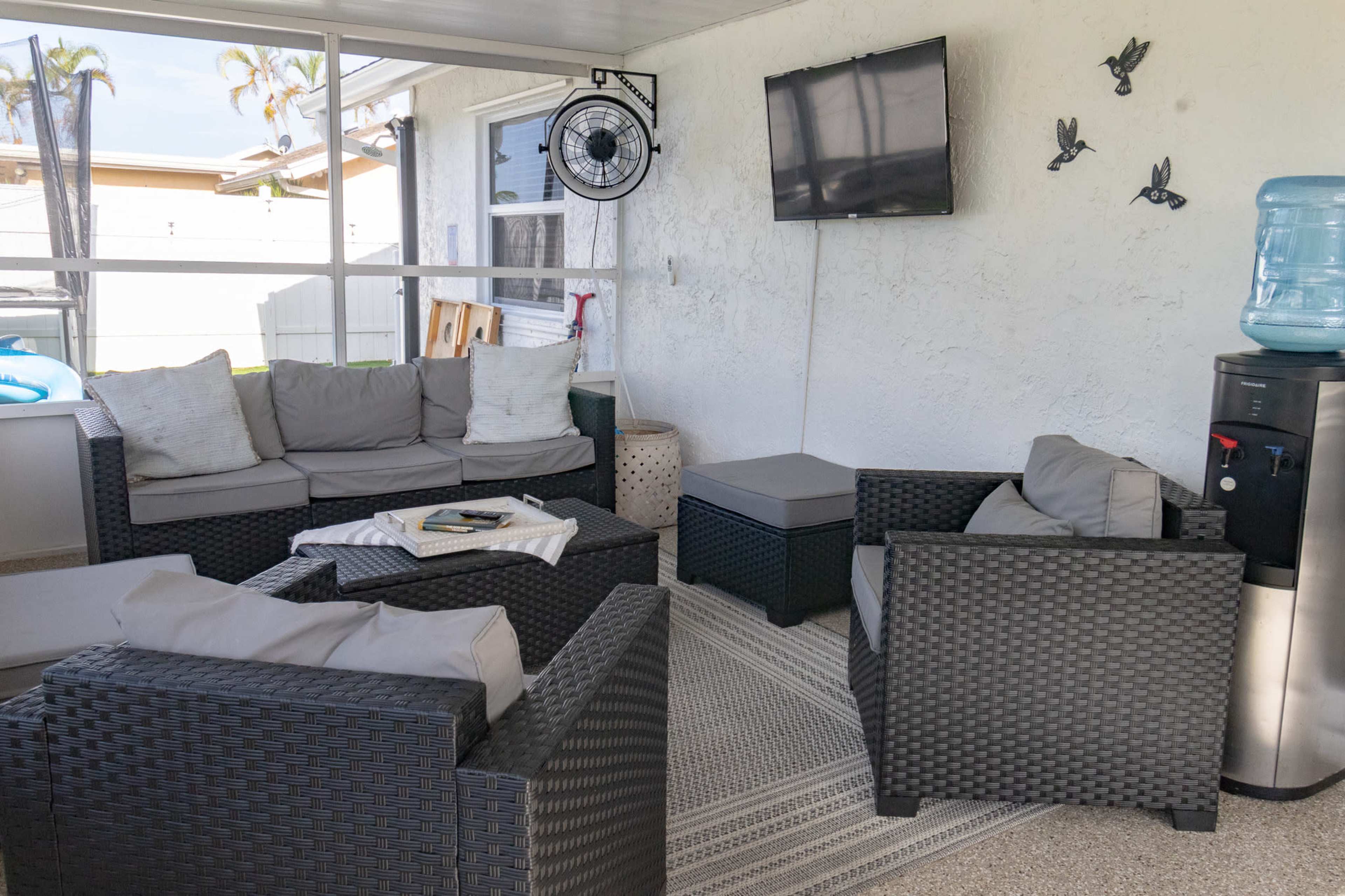 The image shows a screened-in porch area furnished with dark wicker sofas and a coffee table, accompanied by a water cooler and a television mounted on the wall.