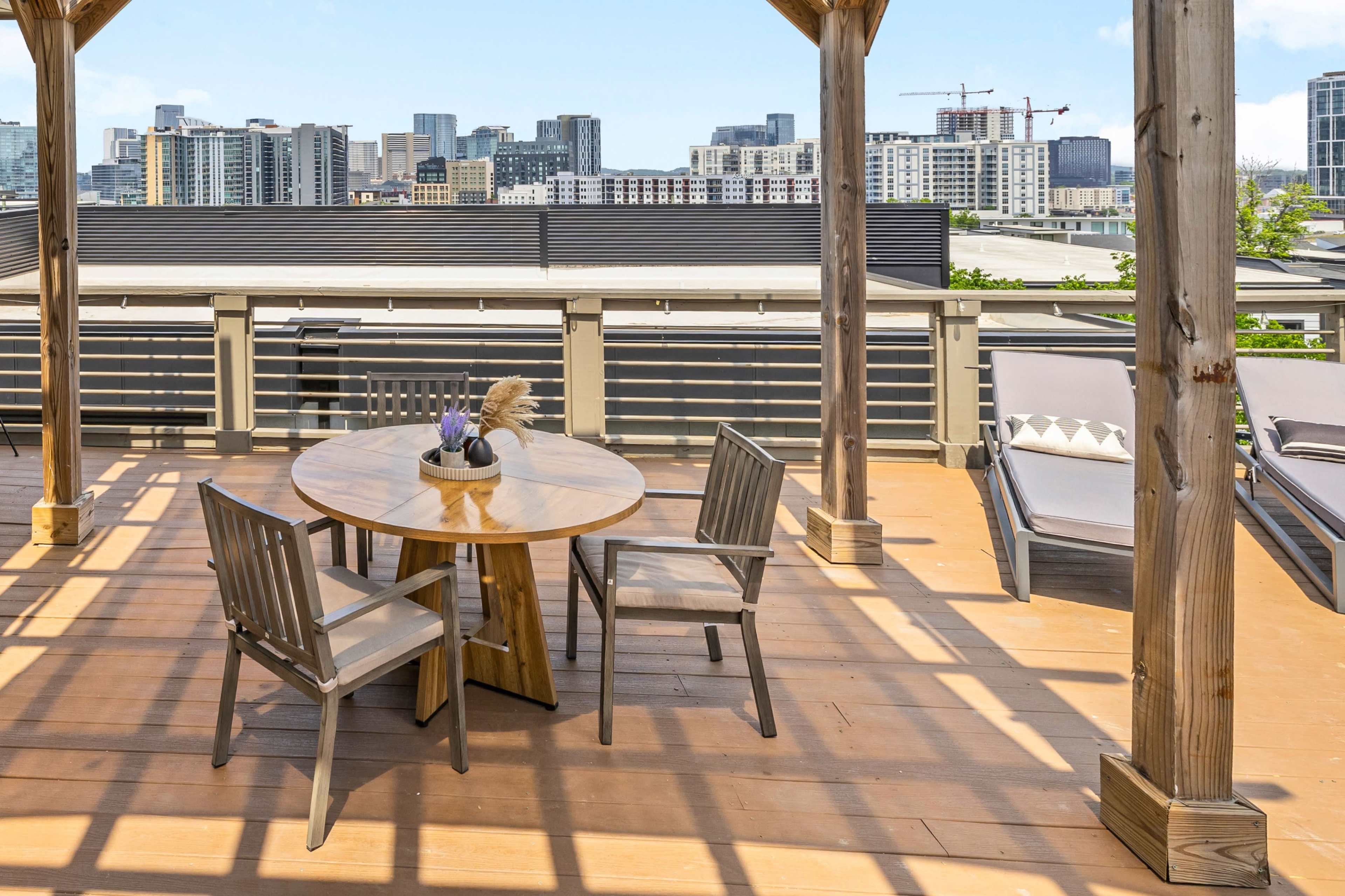 A wooden terrace features a round table with four chairs and a background view of a city skyline with various buildings and cranes.