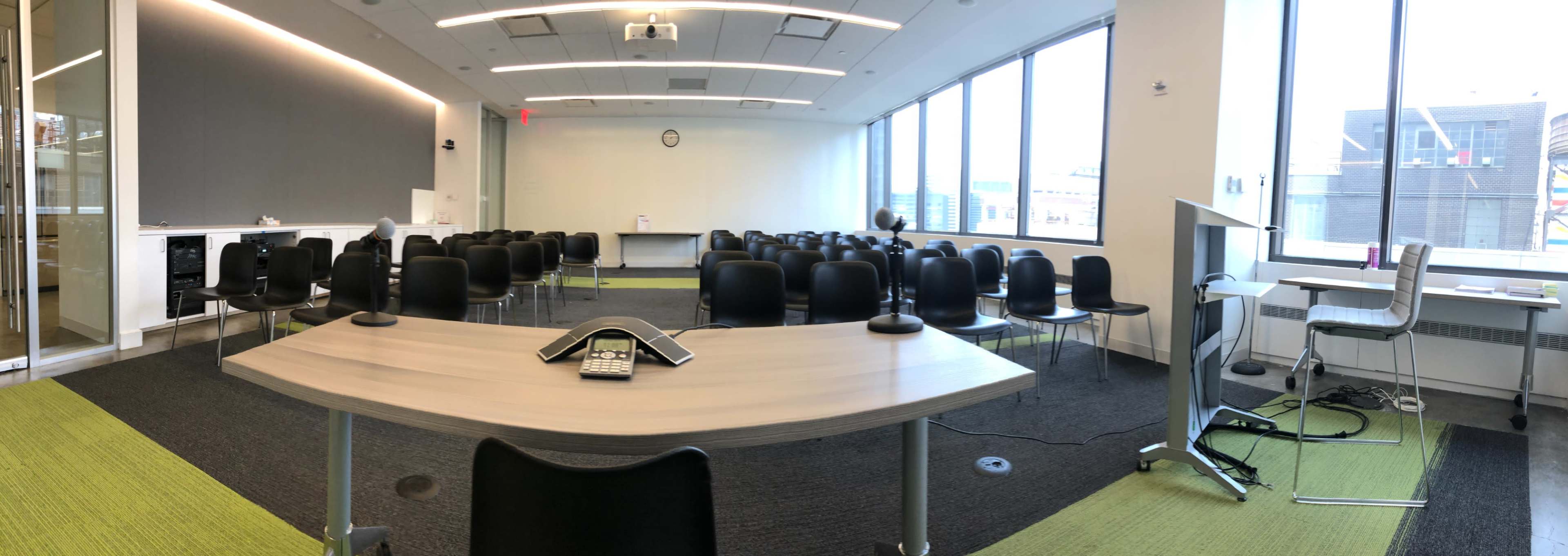 A conference room features a long table with a phone at the center and rows of black chairs set up for a gathering.