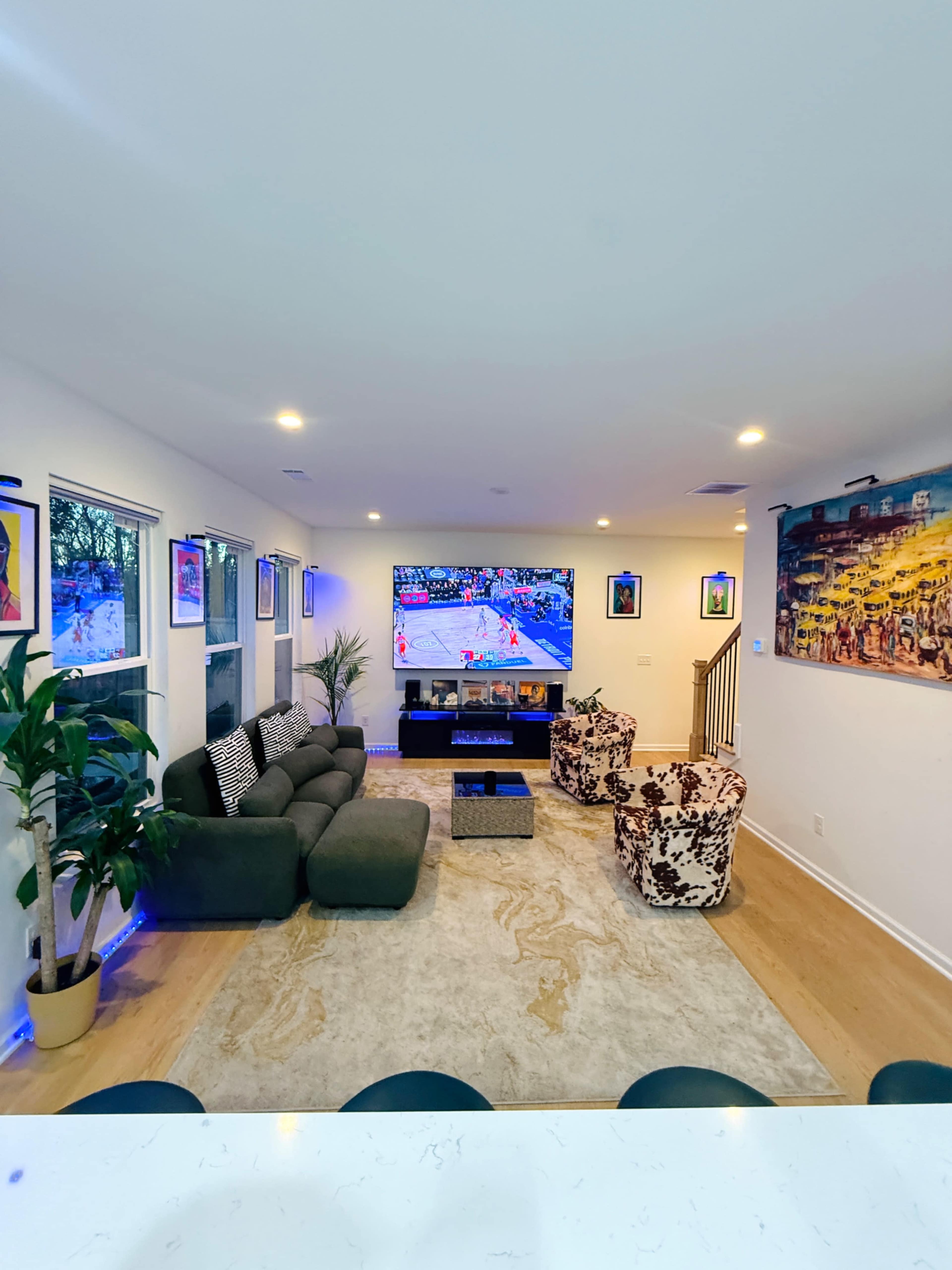 A modern living room features a large television displaying a sports game, surrounded by a sectional sofa, decorative armchairs, and artwork on the walls.