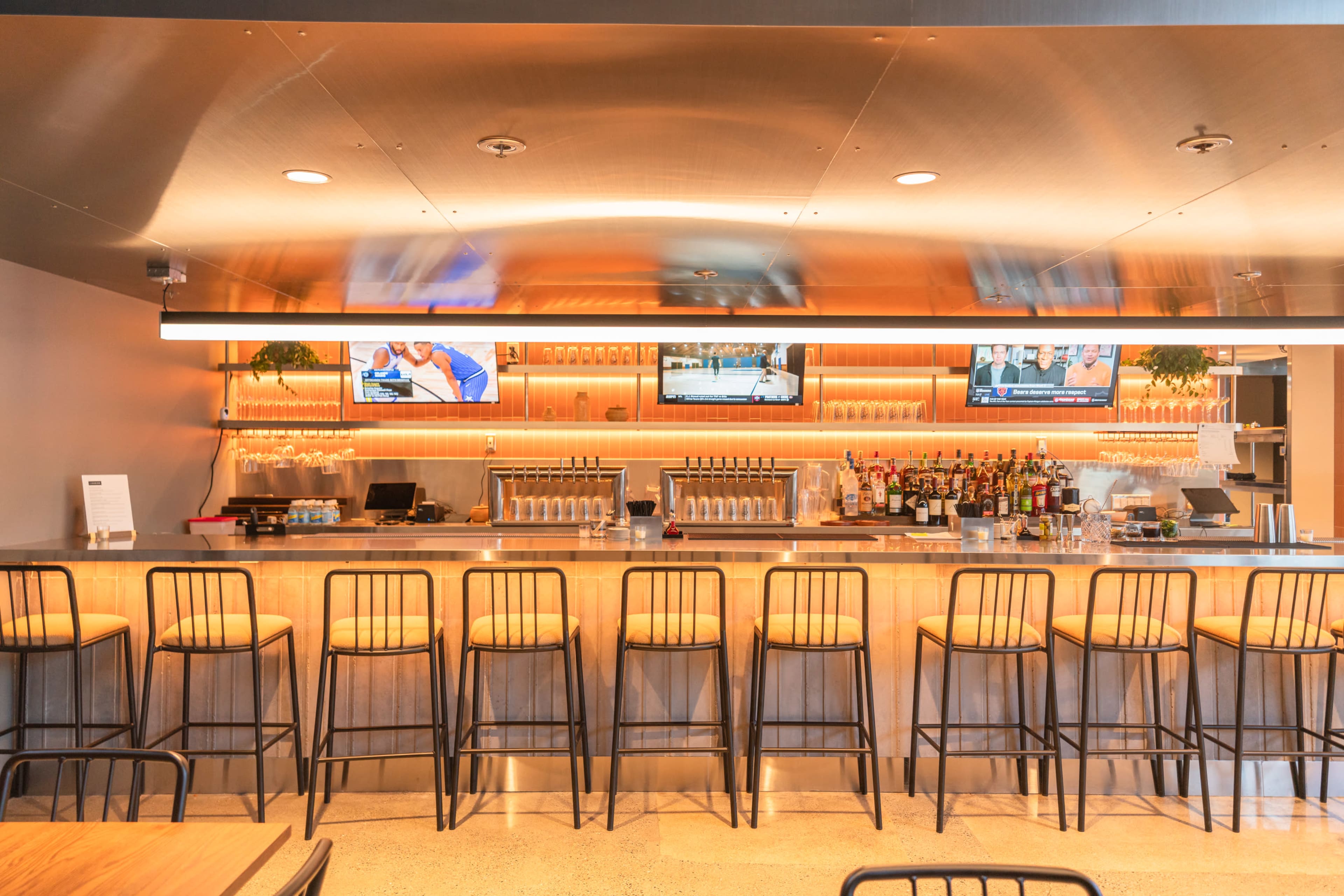 The image shows a modern bar area with a sleek counter, several bar stools, and televisions displaying sports events mounted above the backlit shelves filled with various bottles.