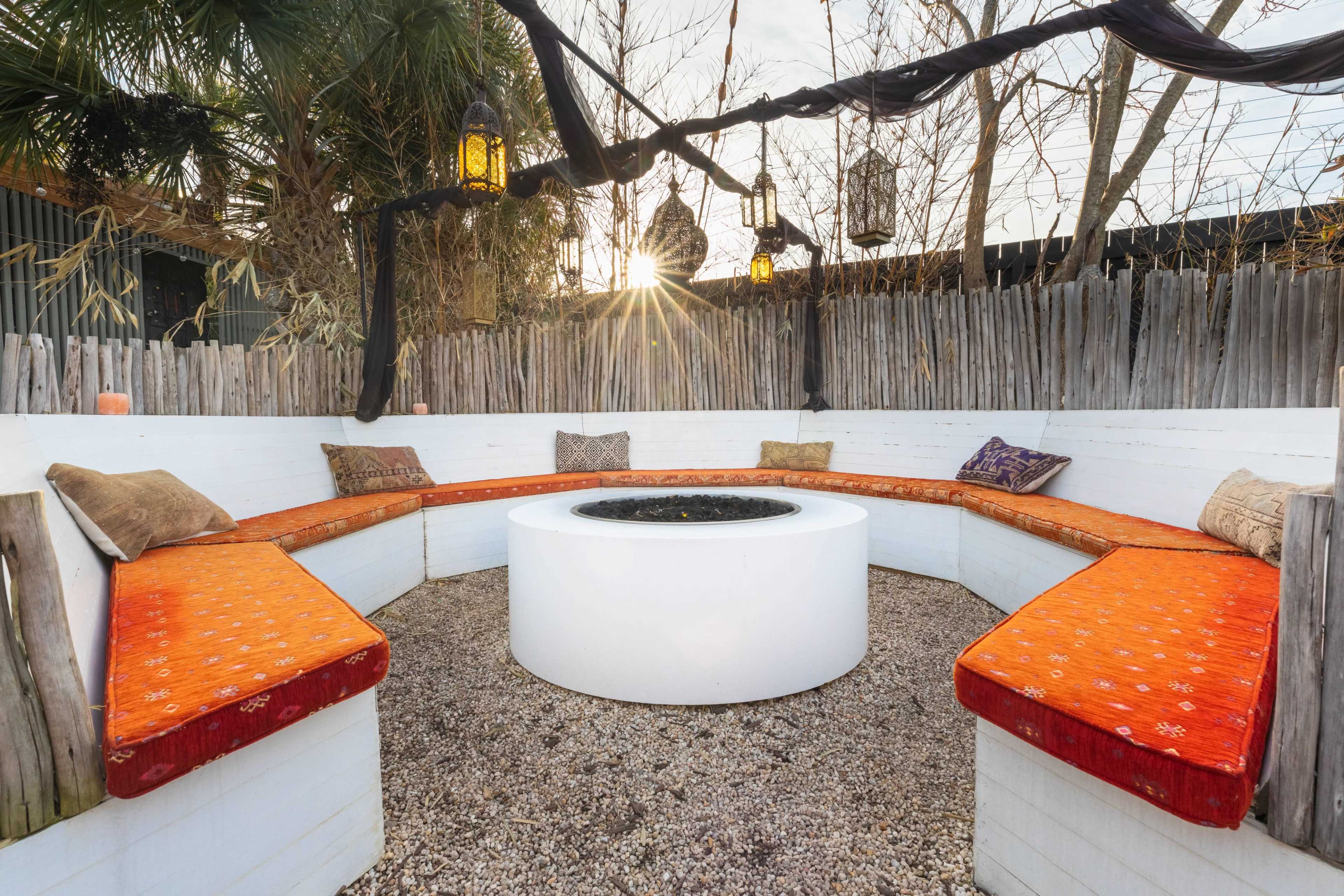 A circular seating area with orange cushions surrounds a central fire pit, set against a backdrop of wooden fencing and hanging lanterns, with a sunset in the background.