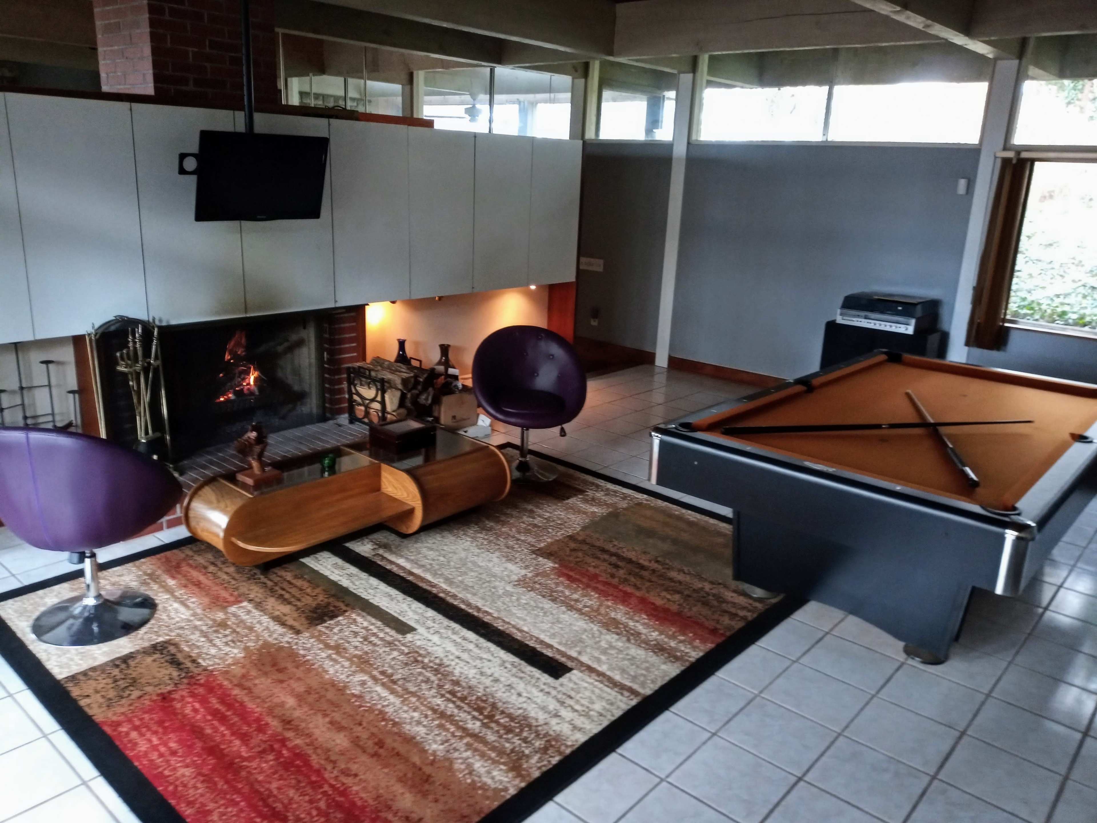 A game room featuring a pool table, a contemporary fireplace with a mounted TV, and modern purple chairs arranged on a patterned rug.
