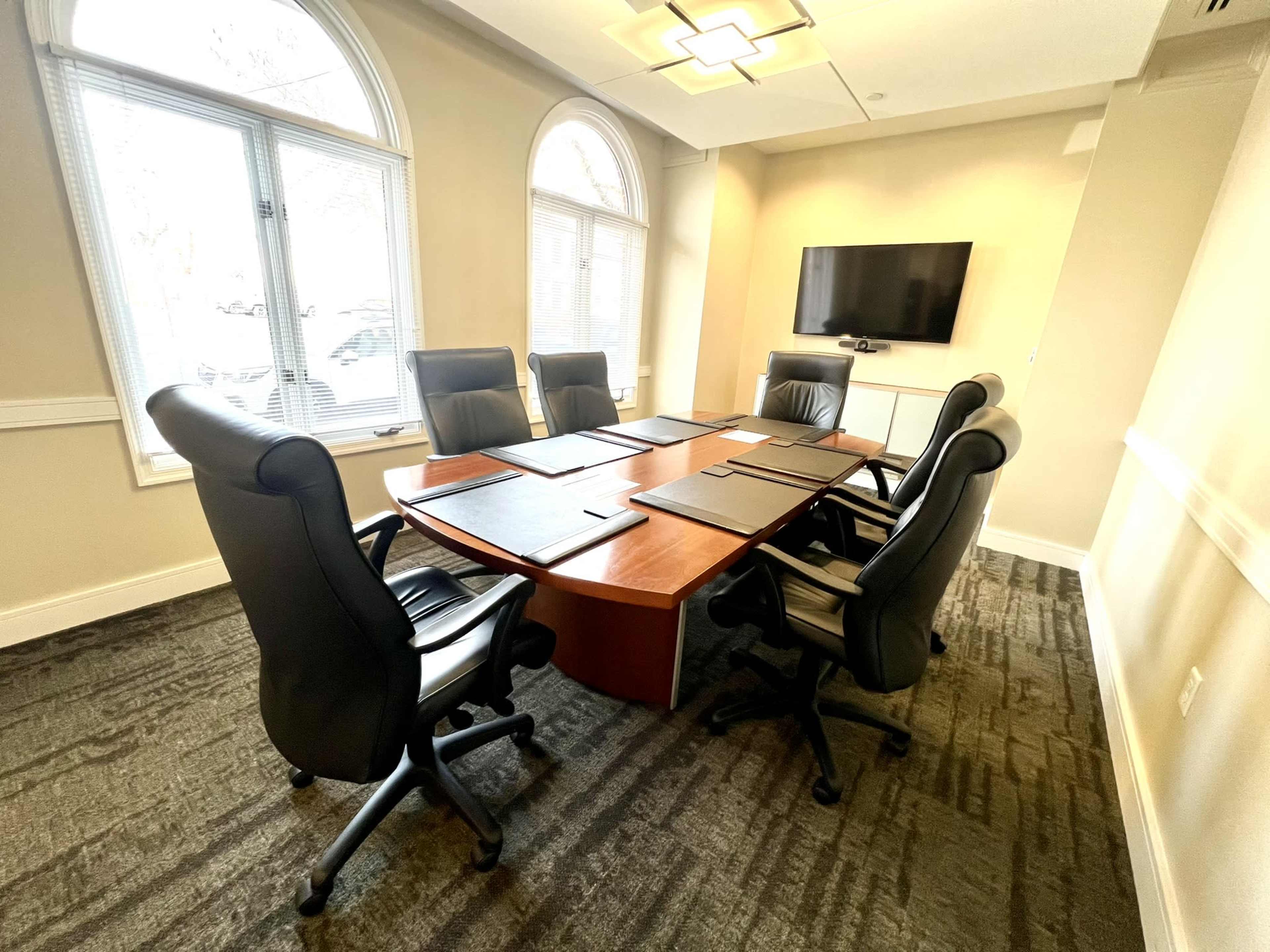 A conference room with a large wooden table surrounded by six black leather chairs and a wall-mounted television.