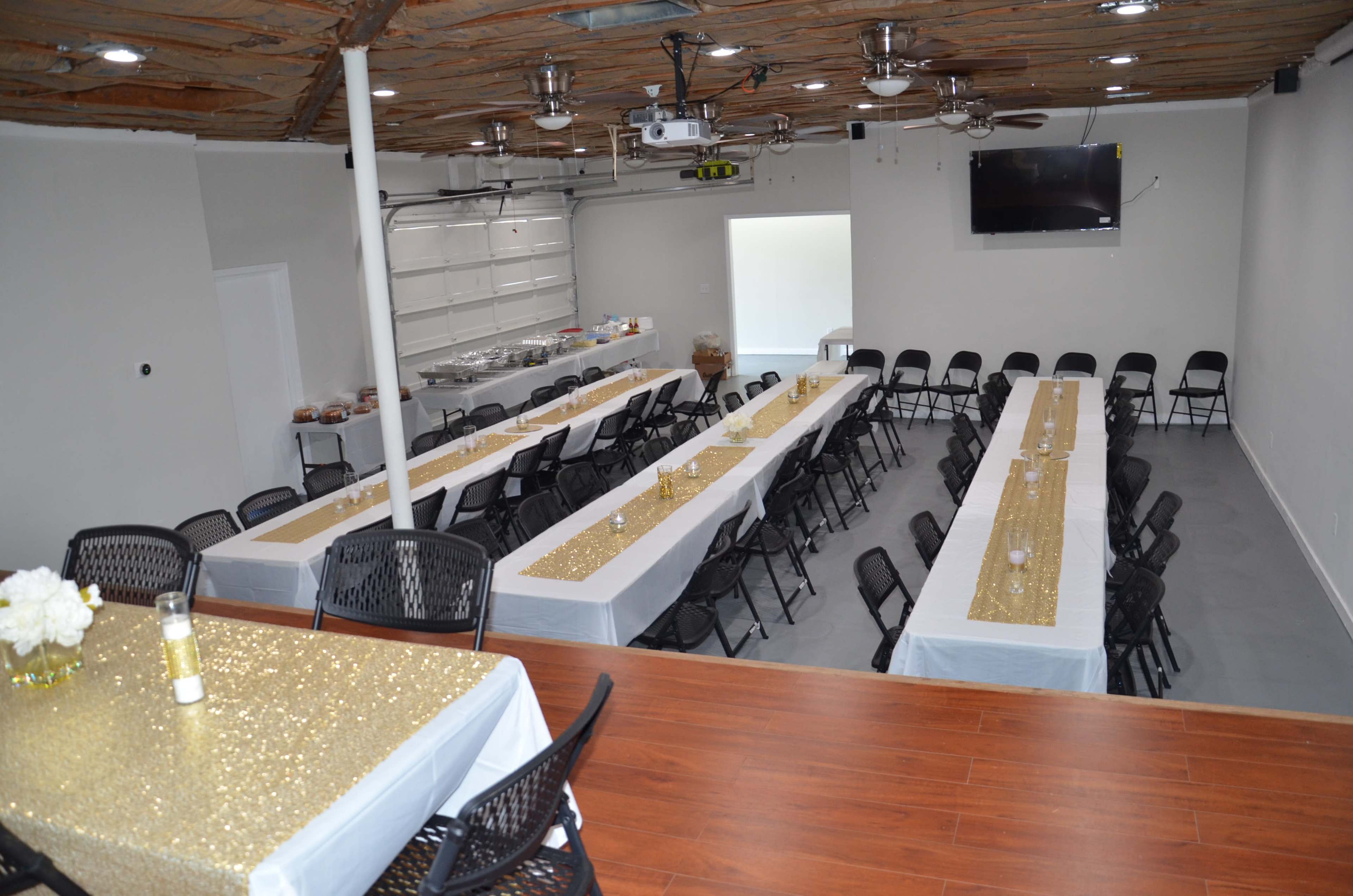 A spacious event room set with multiple long tables covered in white and gold tablecloths, surrounded by black chairs.