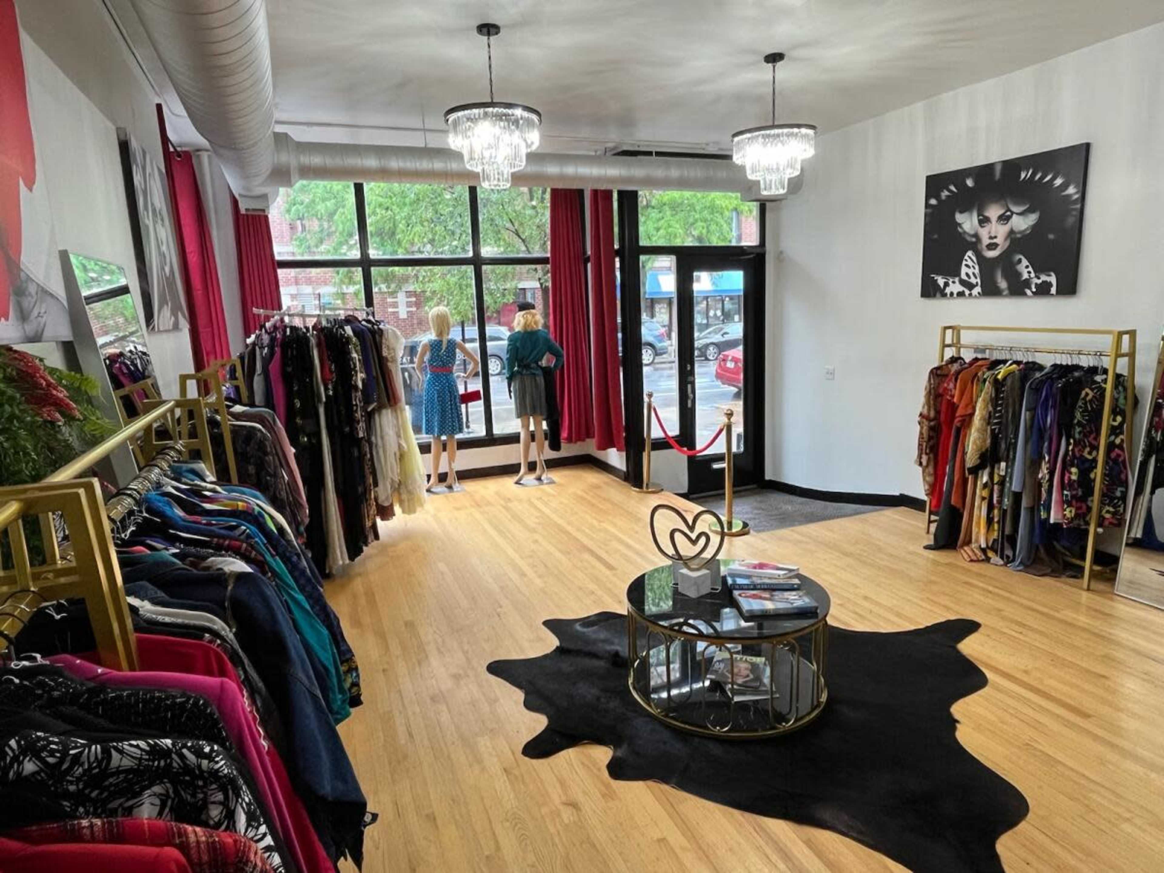 The interior of a clothing store features racks of colorful garments, a round glass table, and two customers looking out through a large front window.