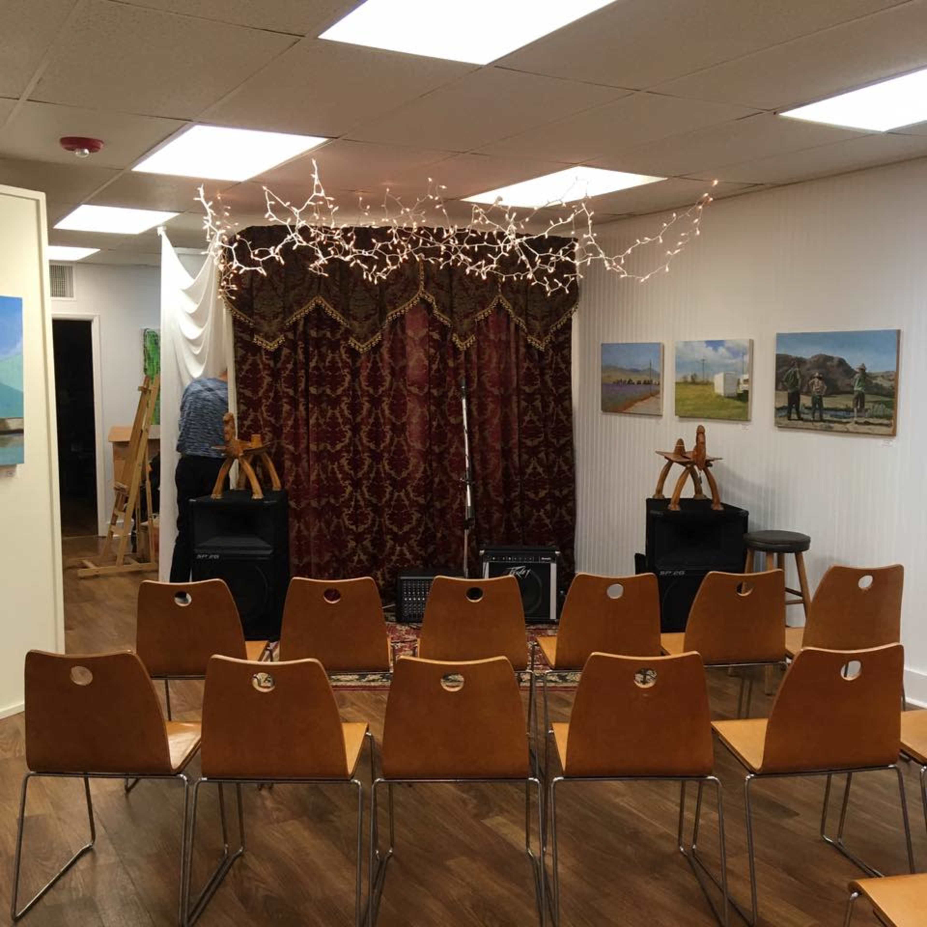 A row of wooden chairs faces a patterned curtain, with art displayed on the walls and decorative string lights overhead.