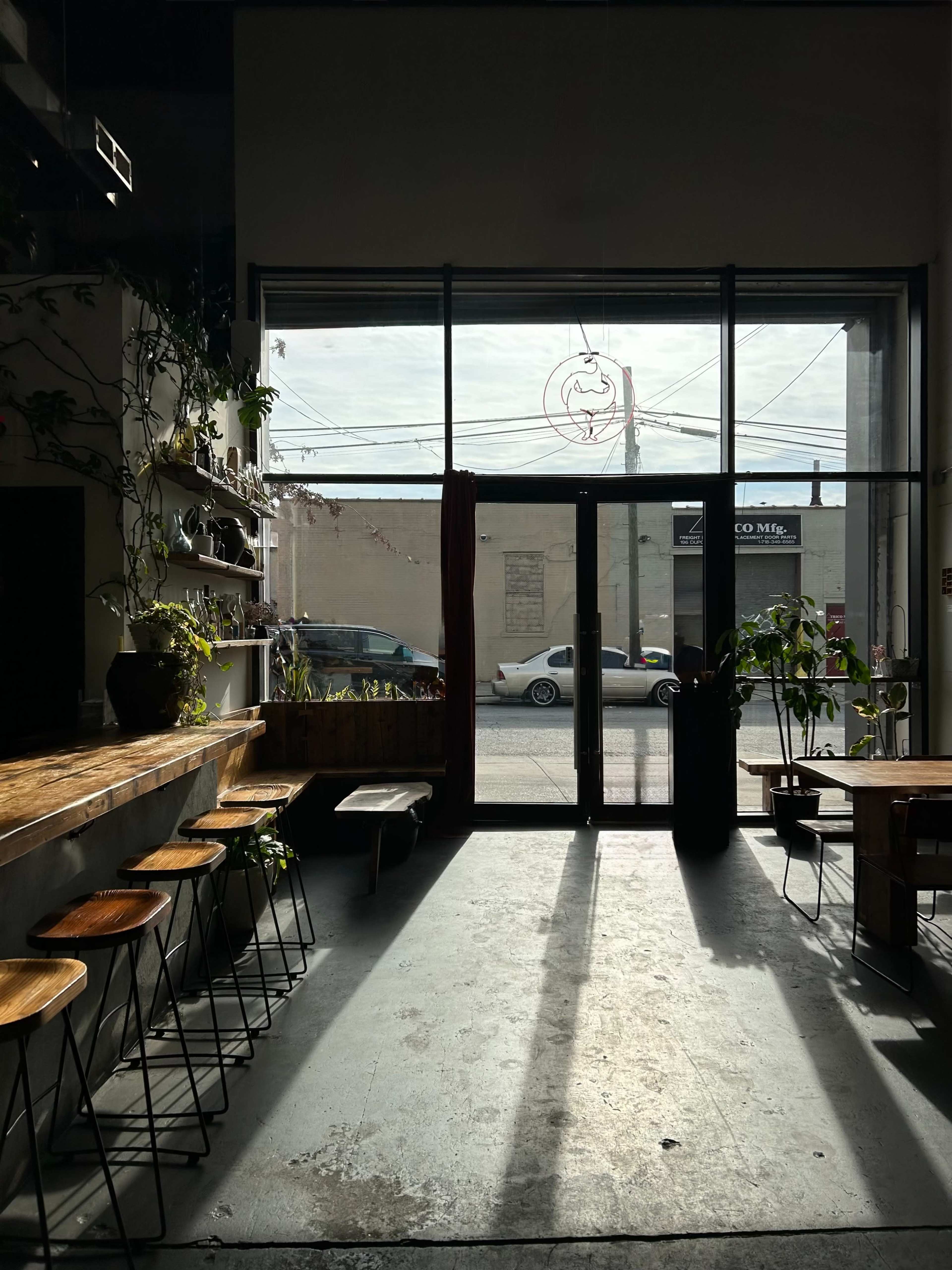 The interior of a café features wooden bar stools along a counter, with large windows allowing natural light and shadows to fill the space.
