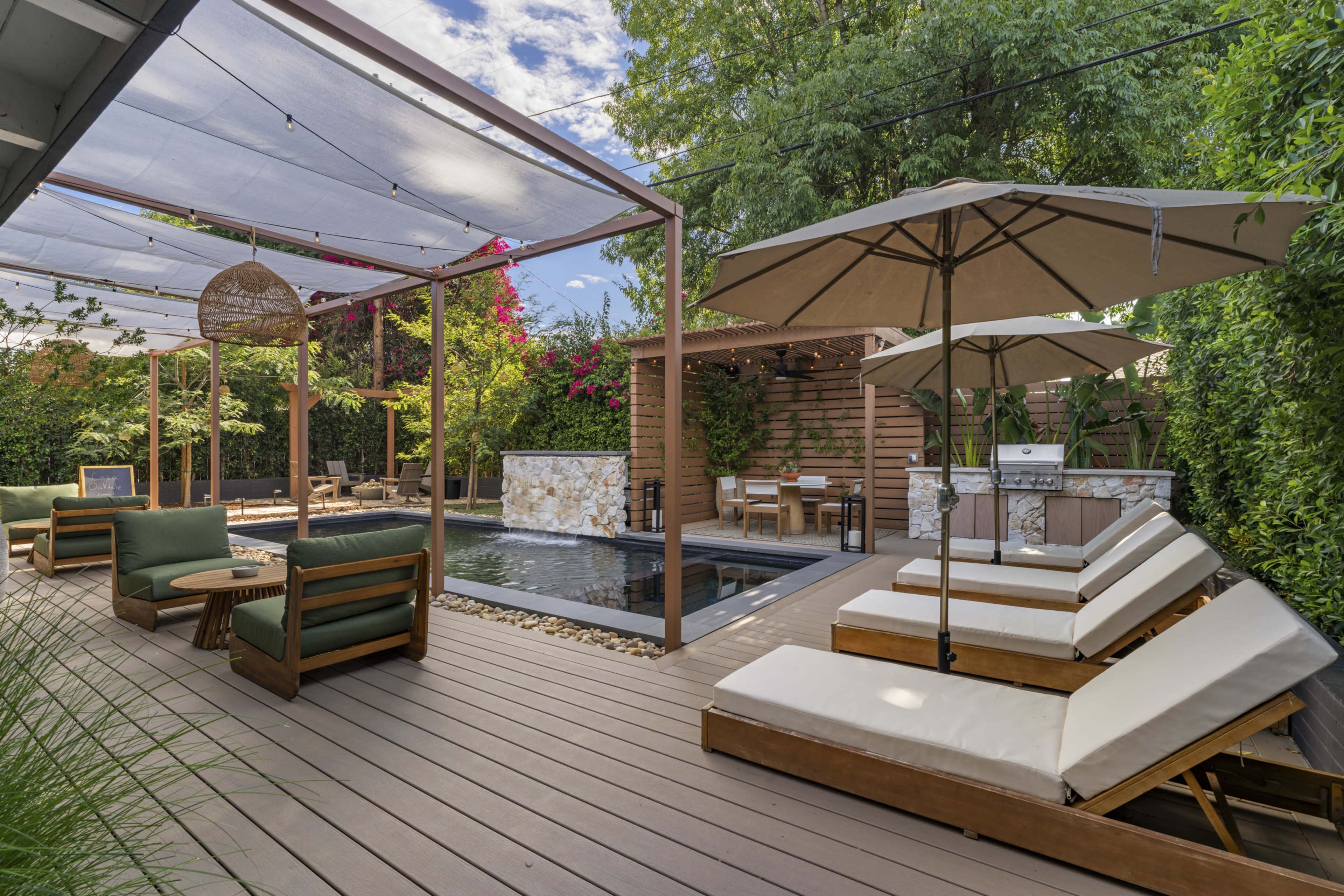 The image shows a landscaped backyard featuring a swimming pool, sun loungers, patio furniture, and shaded dining areas, surrounded by greenery and flowering plants.