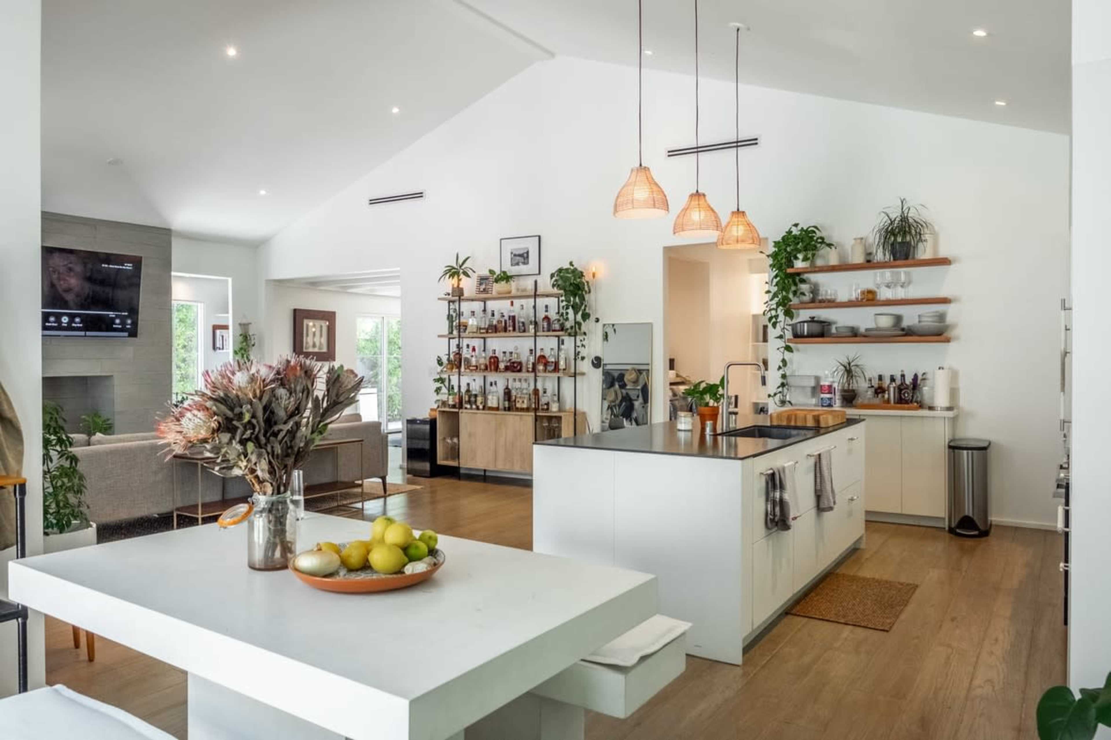 A modern kitchen features an island with a sink, hanging pendant lights, and open shelving filled with bottles and plants, adjacent to a seating area.
