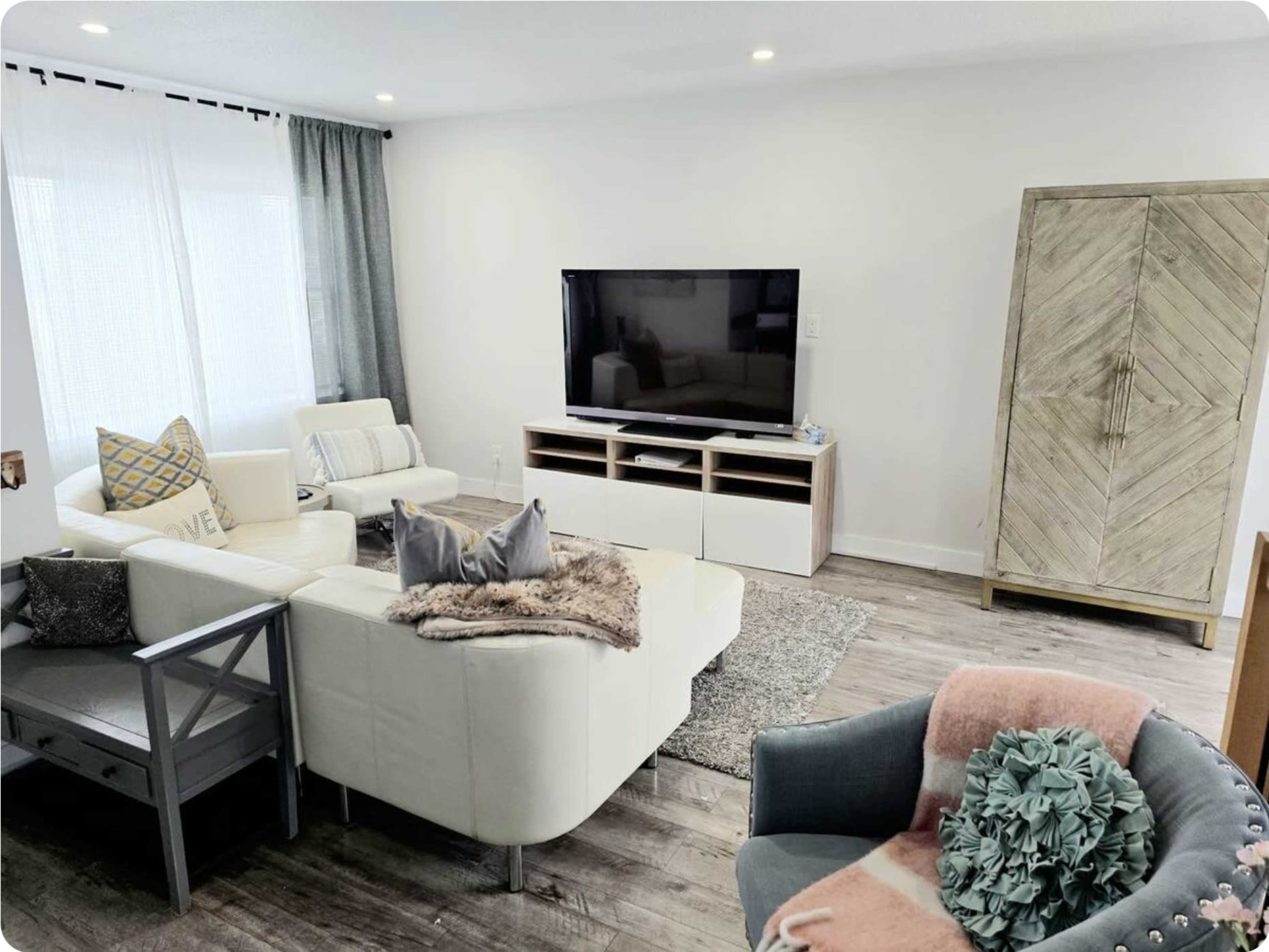 The living room features a white sectional sofa, a large television on a media console, and a wooden cabinet against the wall.