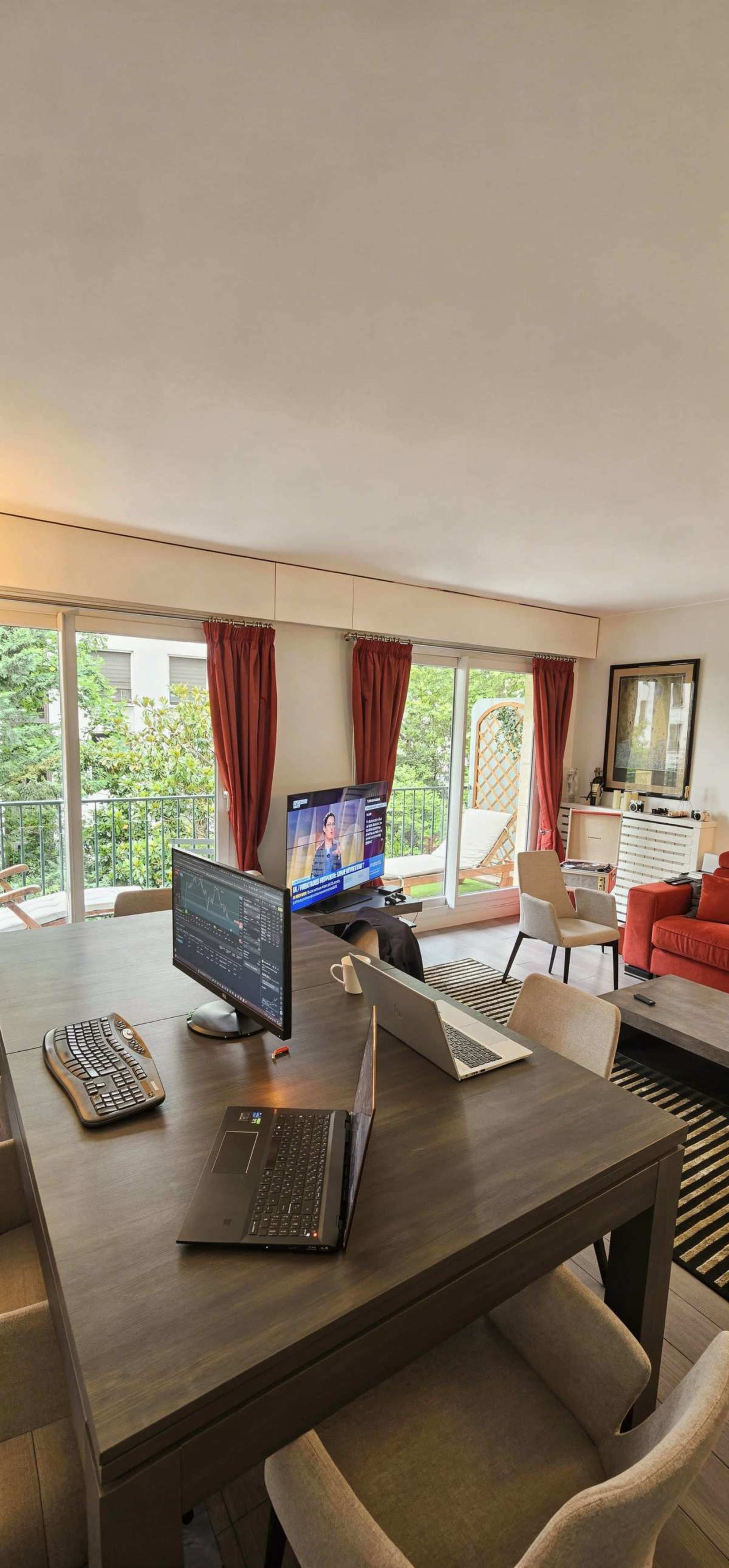 A modern living room features a table with multiple laptops, a television displaying news, and large windows with red curtains overlooking a green outdoor landscape.