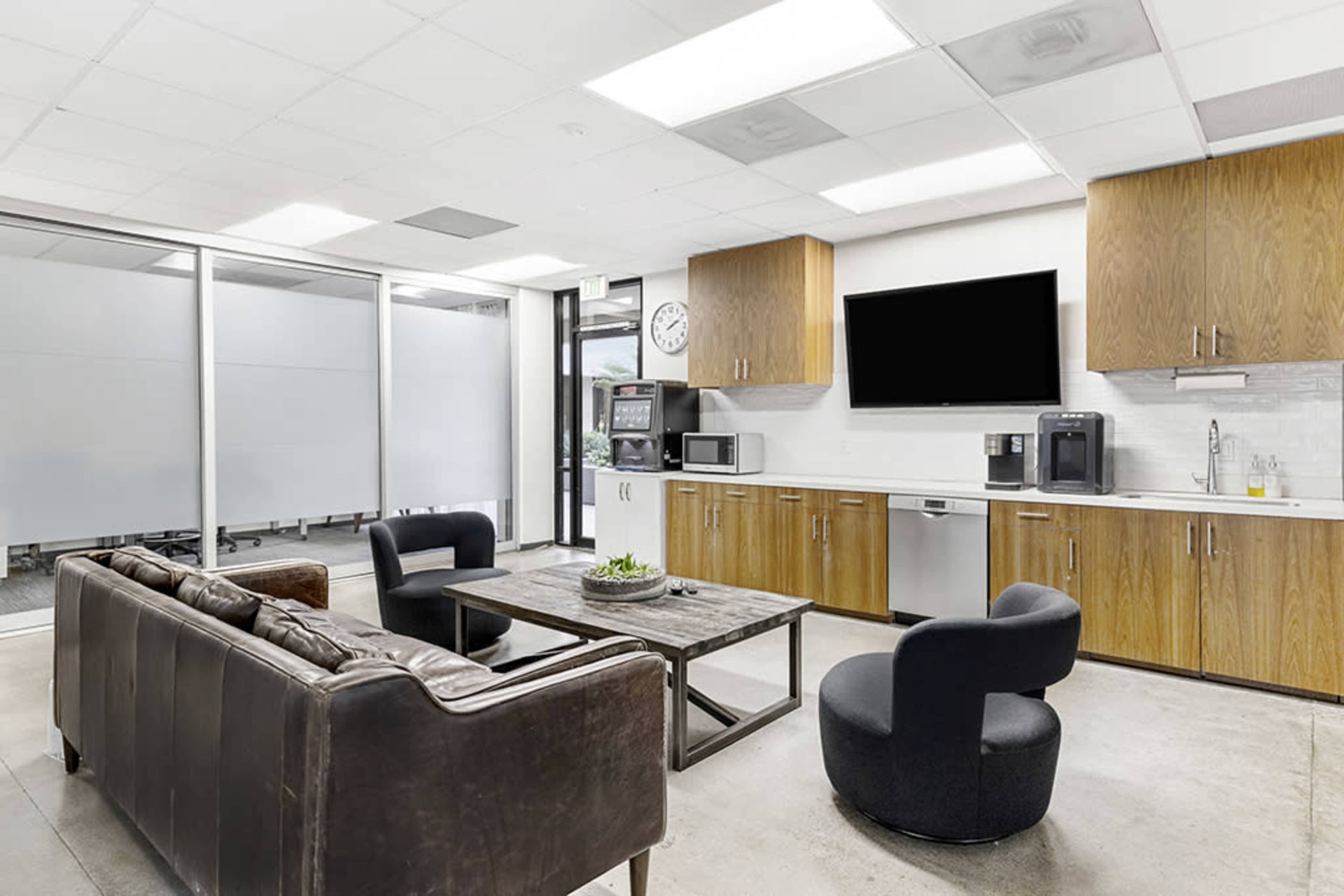 A modern office break room features a seating area with a brown leather sofa, two black chairs, a wooden coffee table, and a kitchenette with appliances and a large wall-mounted television.