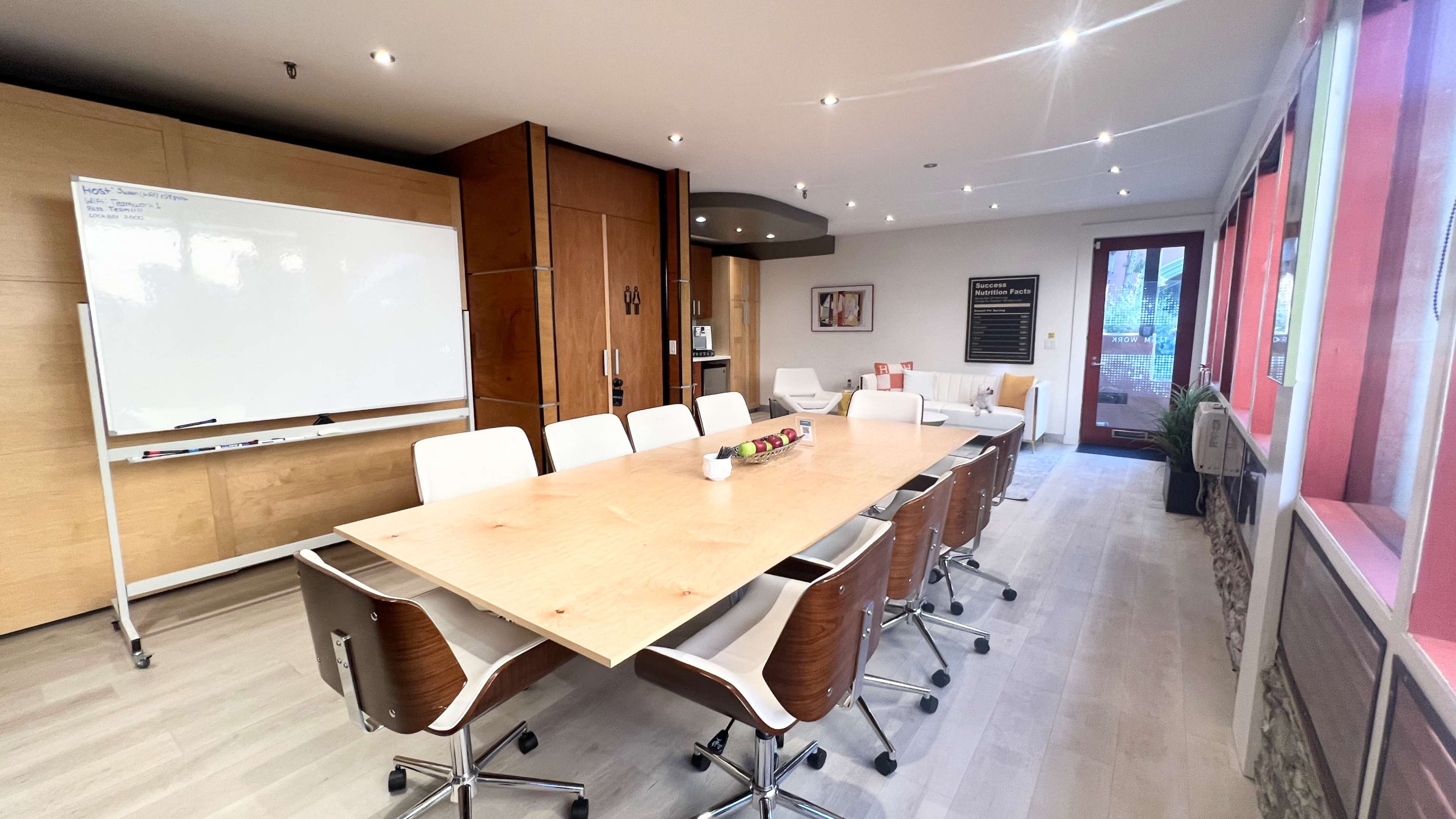A modern conference room with a long wooden table surrounded by white chairs, a whiteboard on the wall, and large windows letting in natural light.
