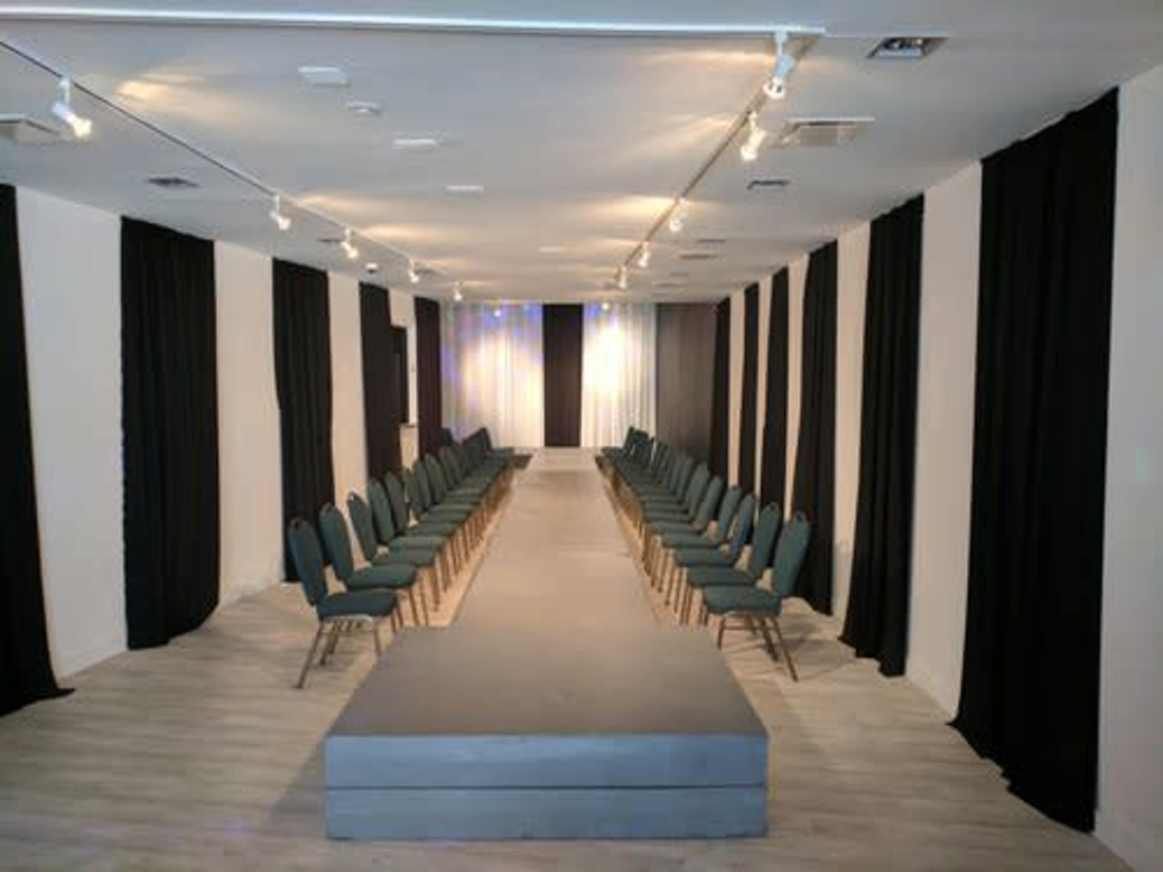 A spacious interior with rows of green chairs on either side of a gray runway, framed by black curtains and soft lighting.