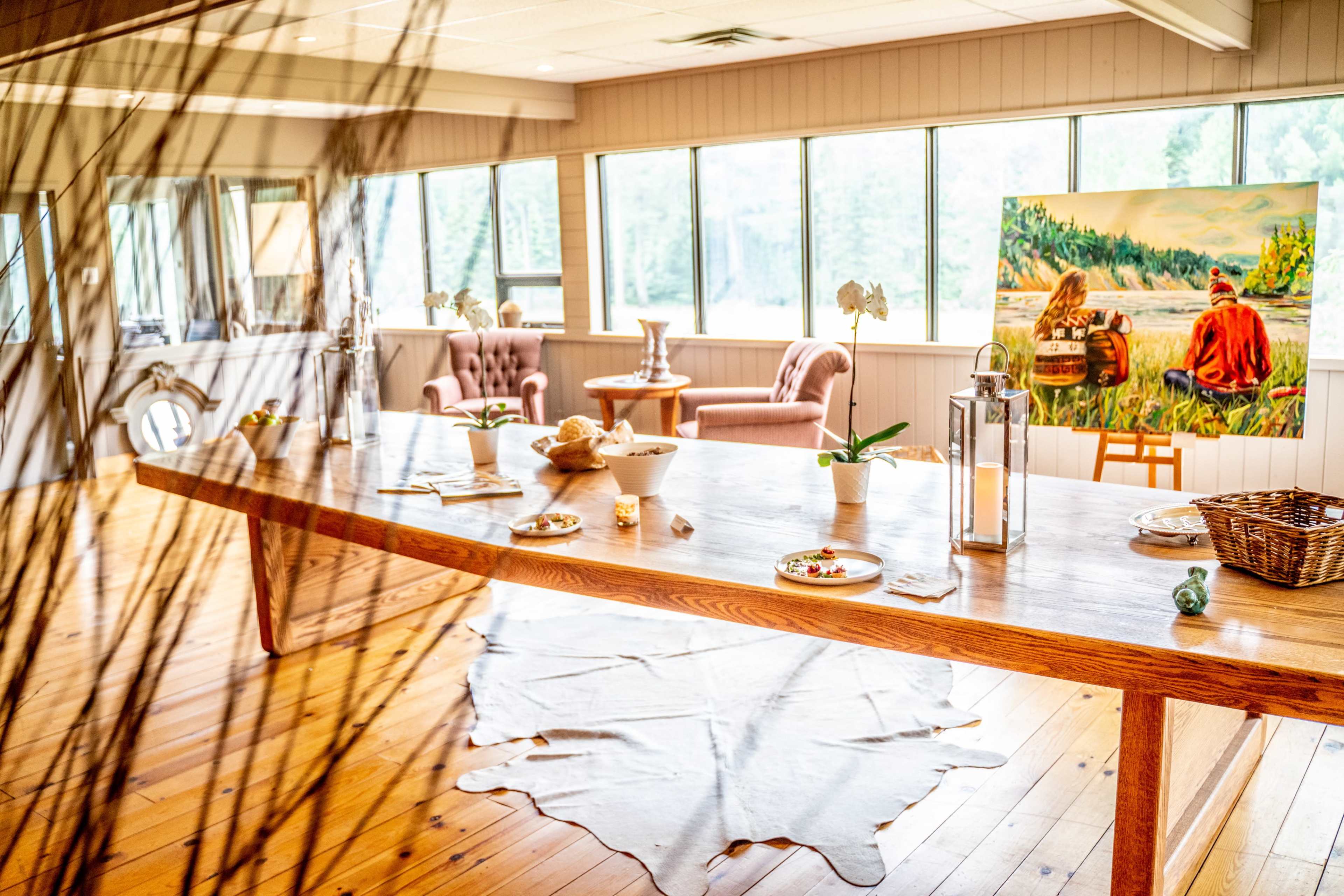 A spacious dining area with a large wooden table set for a meal, surrounded by natural light and featuring a painting on the wall.