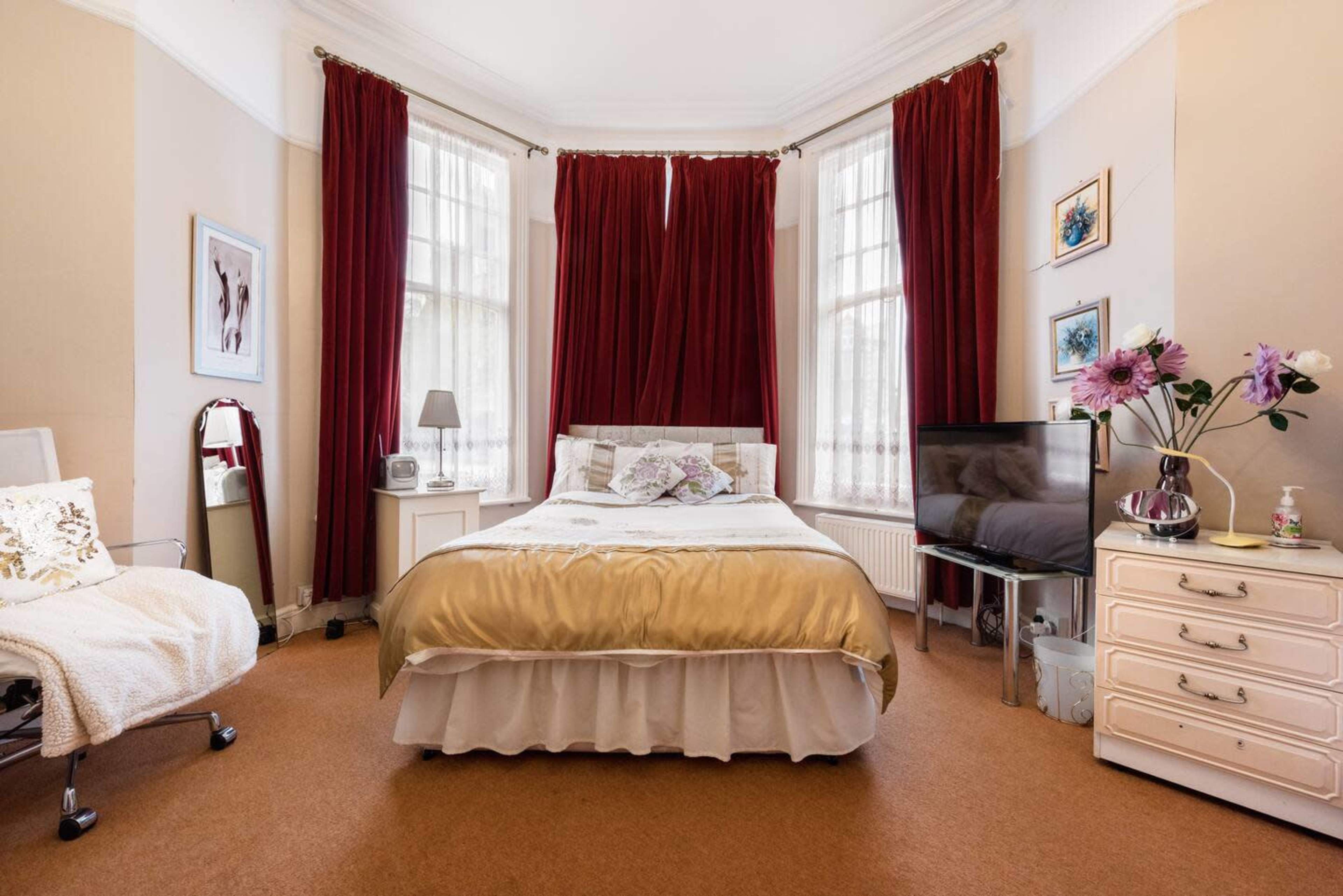 A bedroom features a bed with gold bedding, large windows with red curtains, and a dresser with a television and decorative flowers.