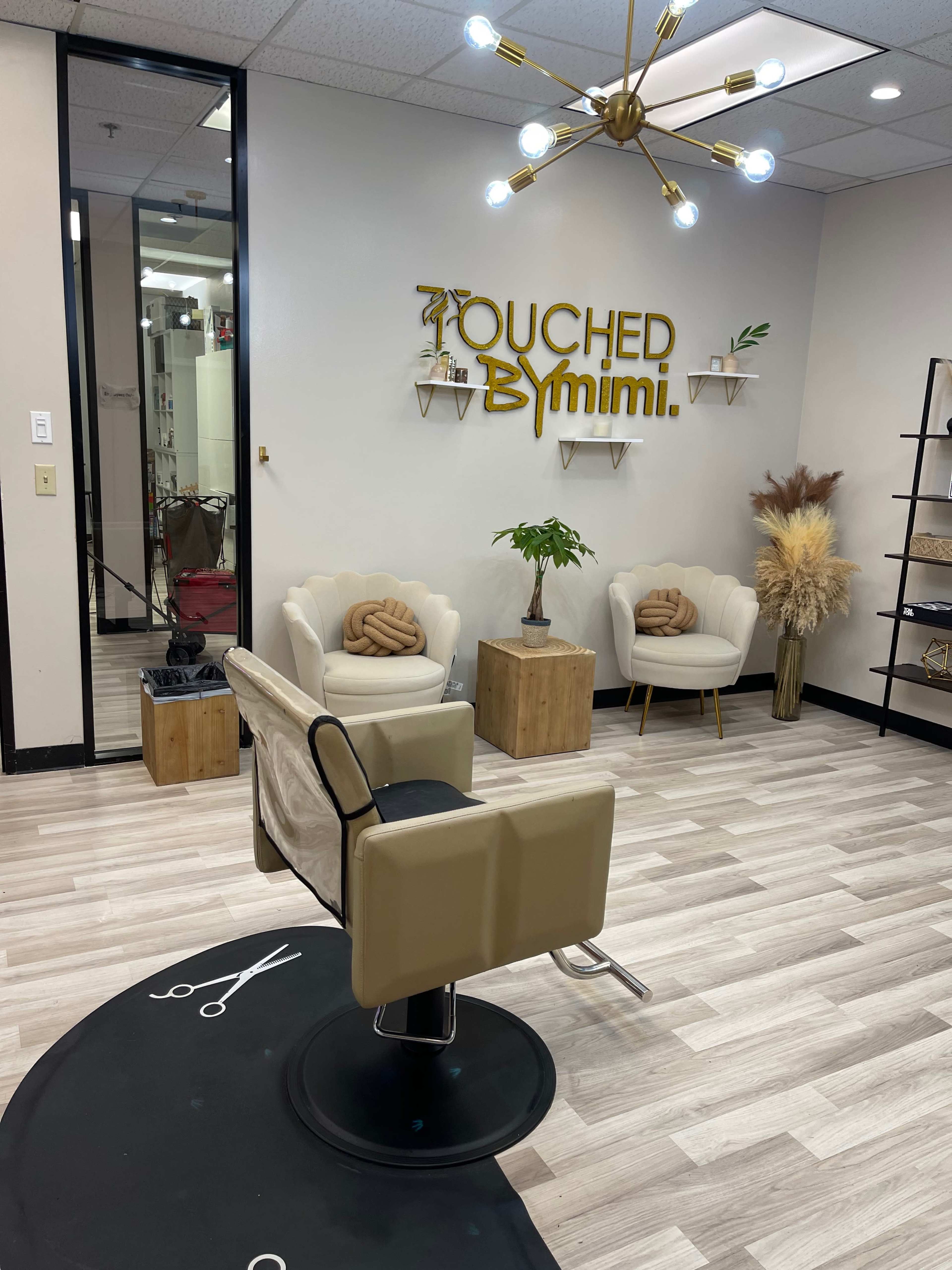 The image shows a salon interior featuring a styling chair, two armchairs, decorative plants, and a wall sign that reads "Touched By Mimi."