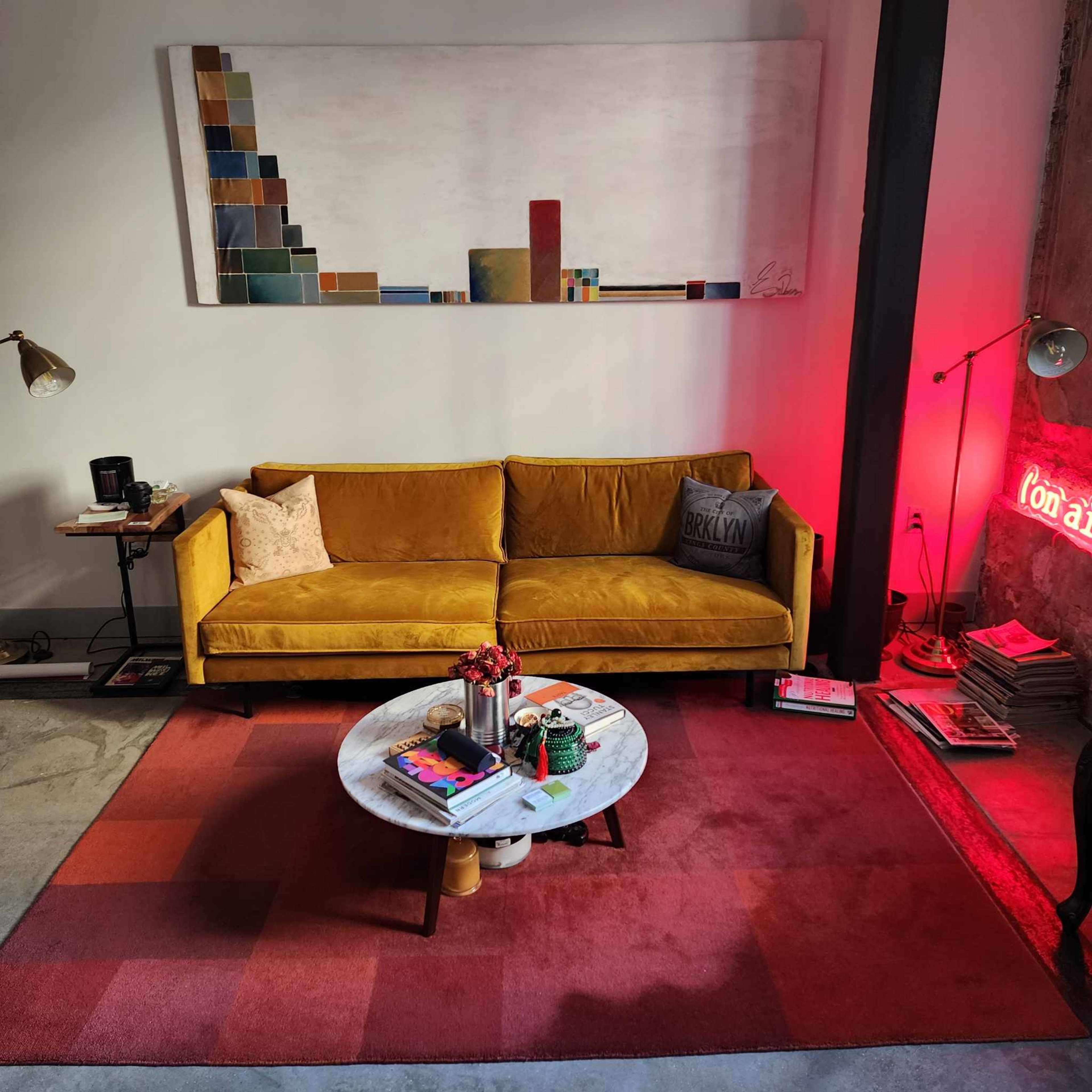 A cozy living room featuring a yellow sofa, a round coffee table with magazines and plants, and colorful artwork on the wall, illuminated by a red neon light.