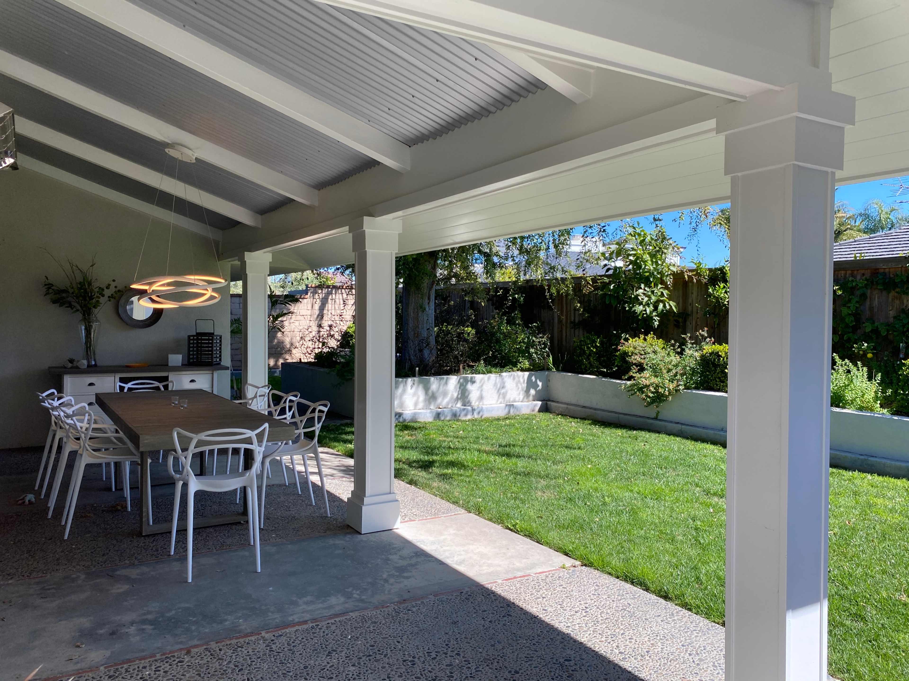 A covered outdoor dining area features a long table surrounded by white chairs, with a grassy lawn and garden visible in the background.
