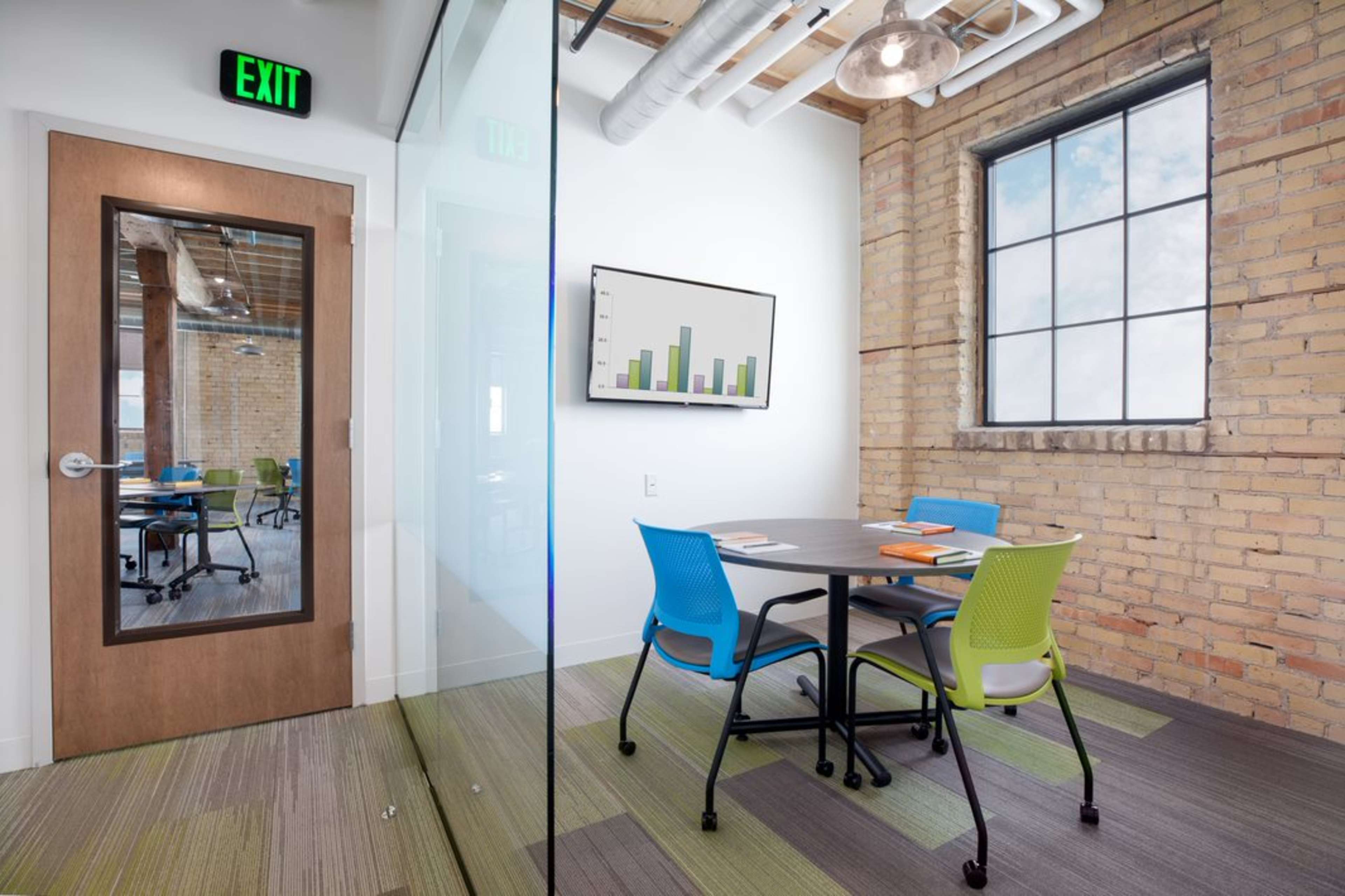 A small meeting room with a round table and colorful chairs, situated near a glass wall and a door marked with an exit sign.