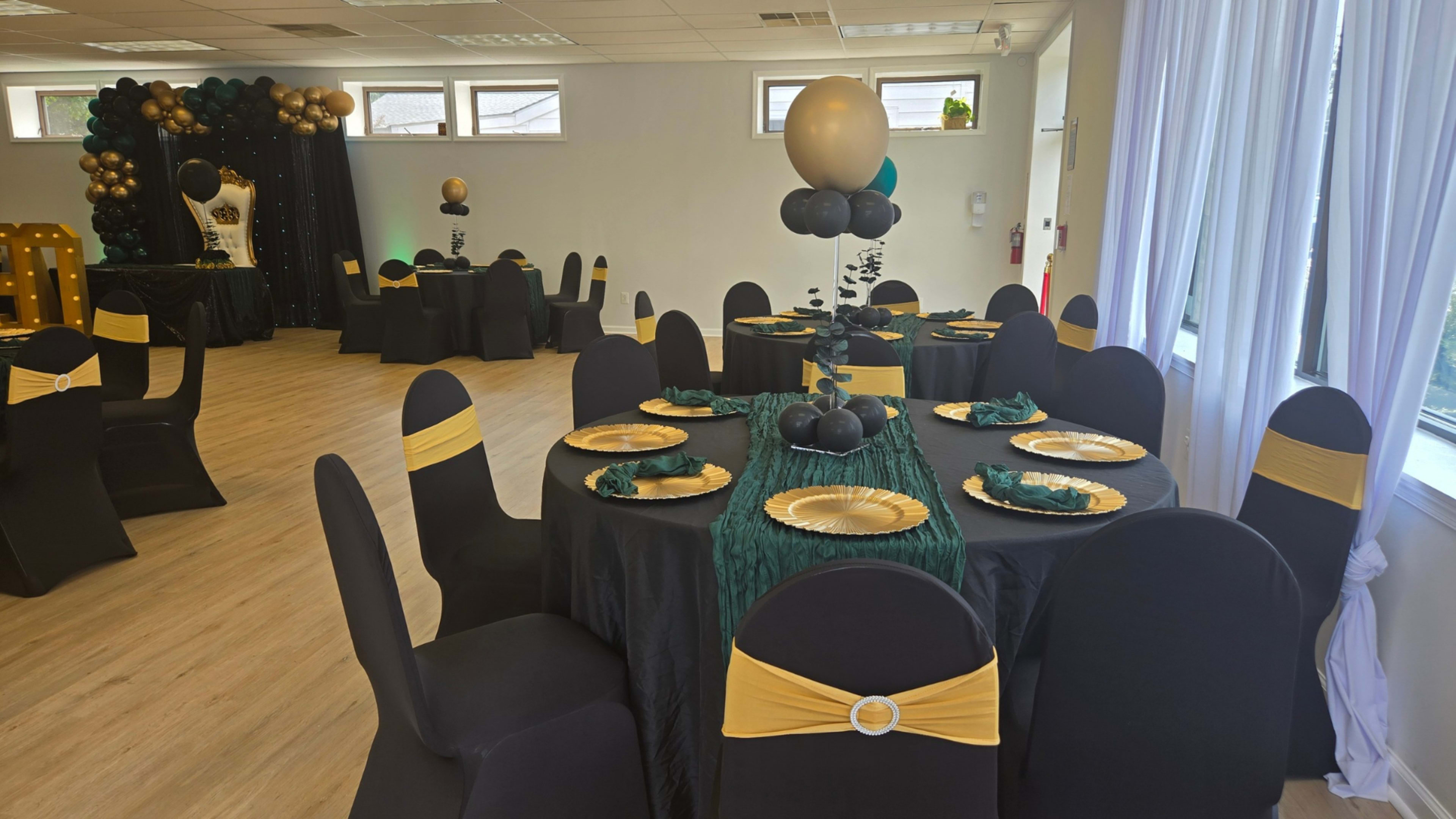The image shows a decorated event space with black and gold tables, chairs with yellow sashes, and a backdrop featuring balloons and lights.