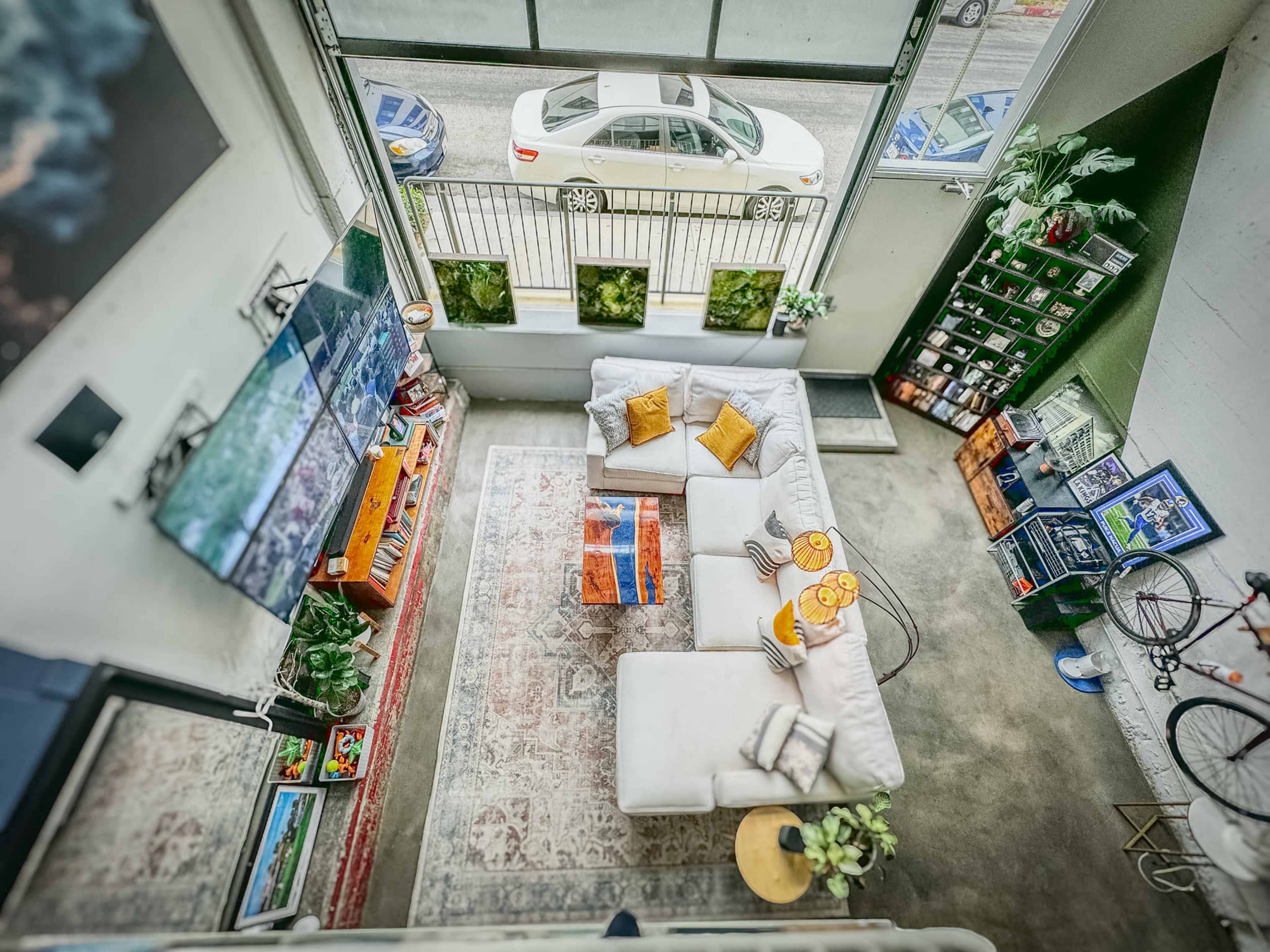A modern living room with a white sectional sofa, a large television mounted on the wall, and a view of a street through a large window.