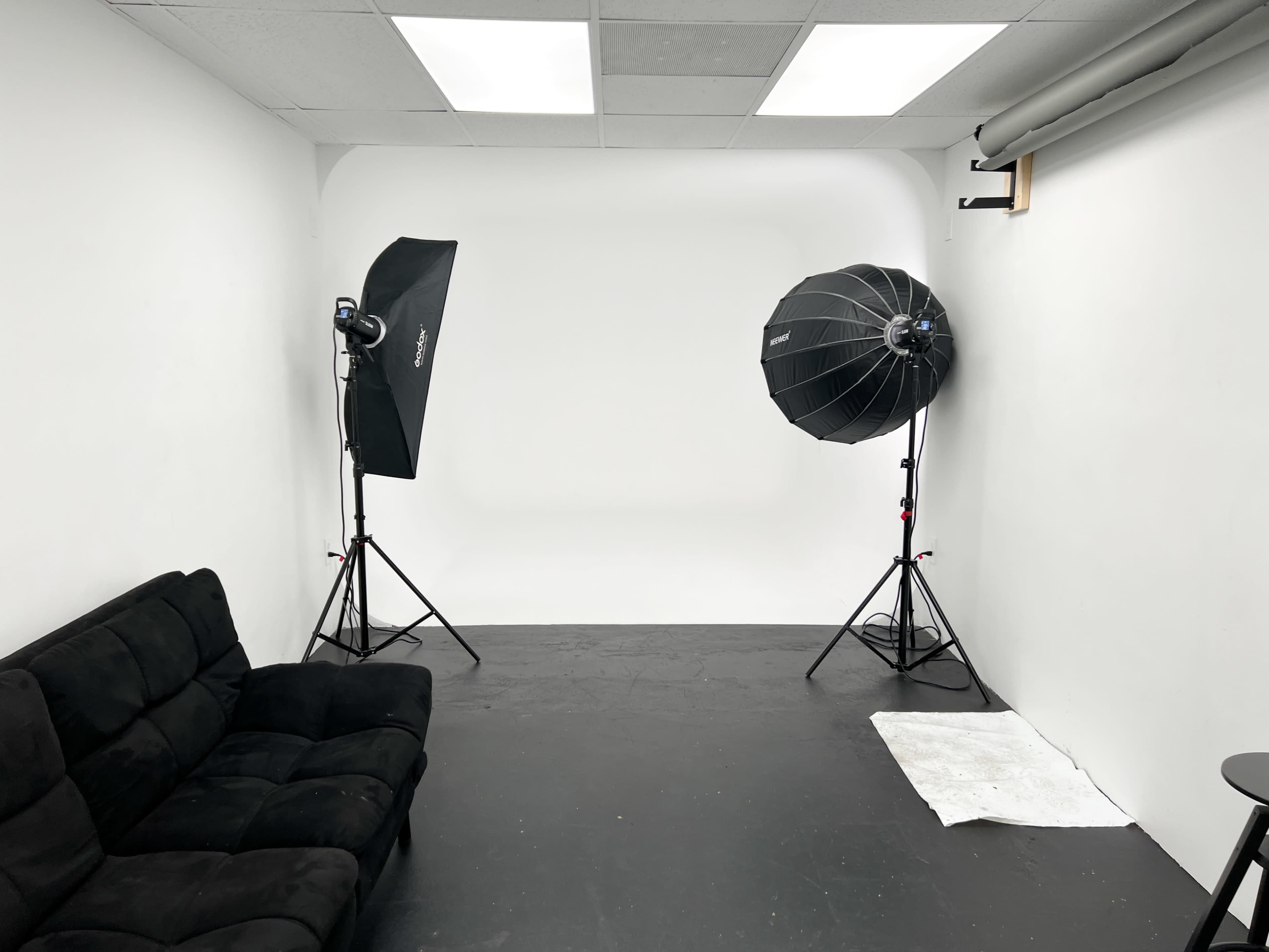 A photo studio features a black couch, two light stands with softboxes, and a white backdrop.