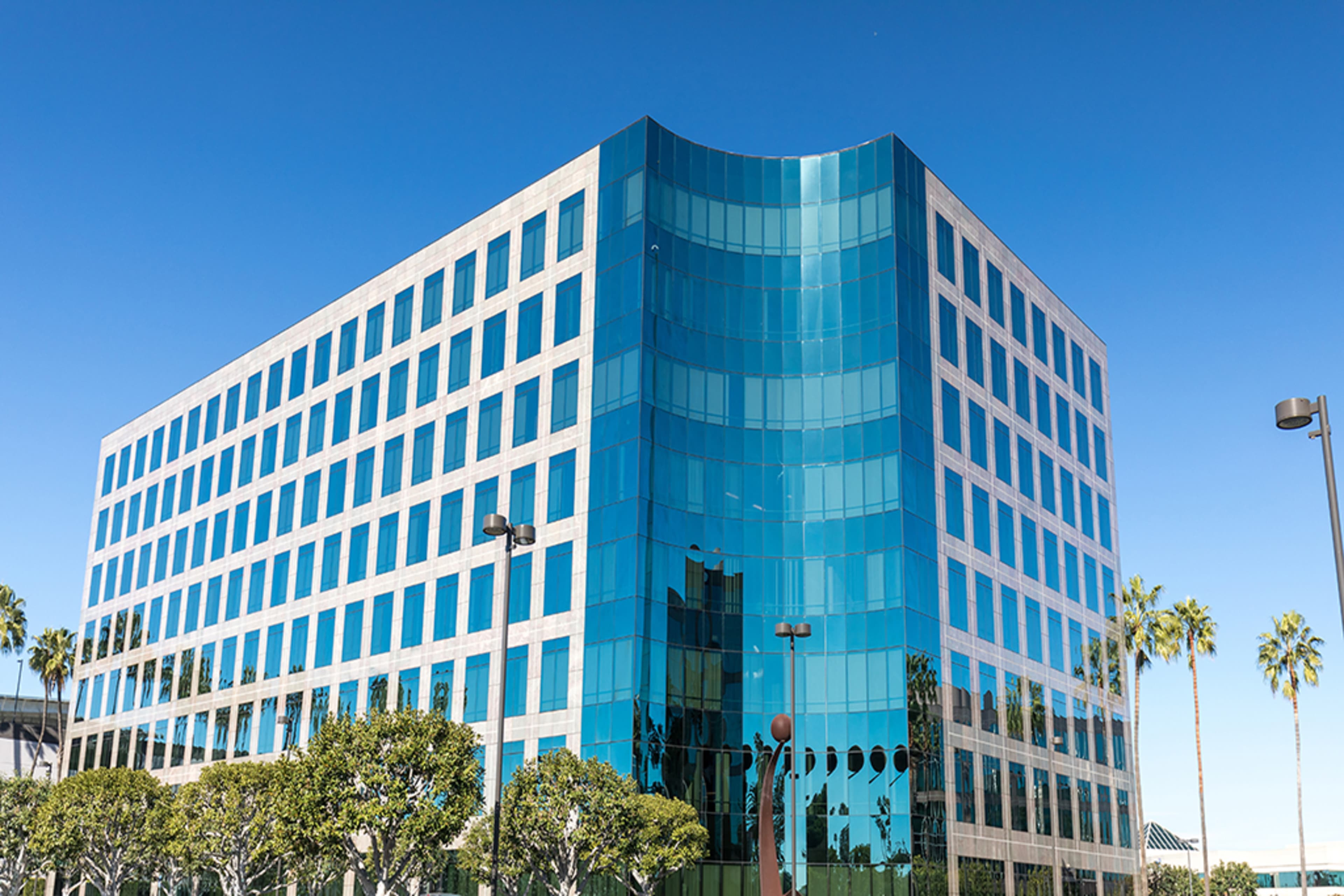 A modern office building features a curved glass facade and is surrounded by palm trees against a clear blue sky.