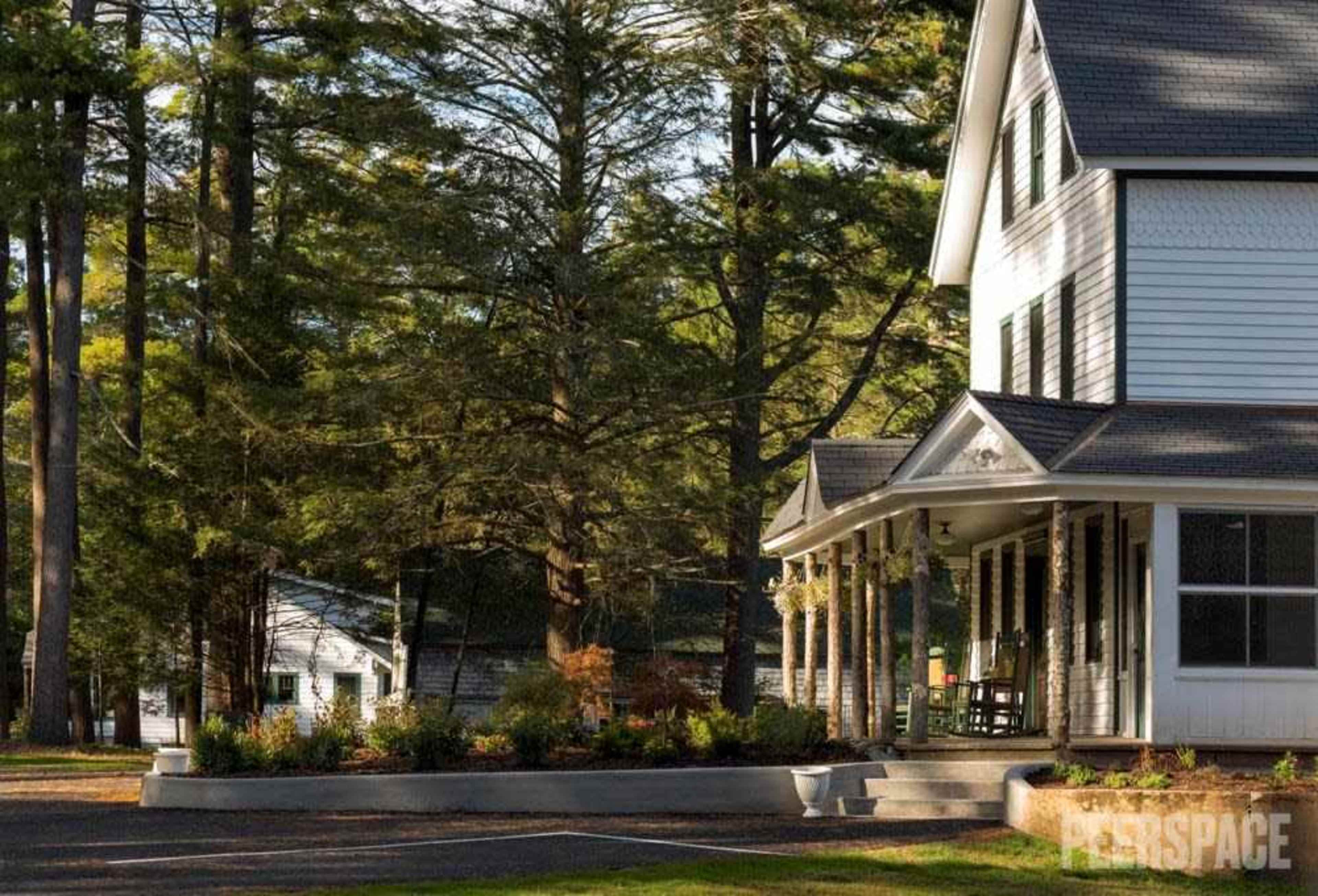 A white two-story house with a porch is surrounded by tall trees and a landscaped garden.
