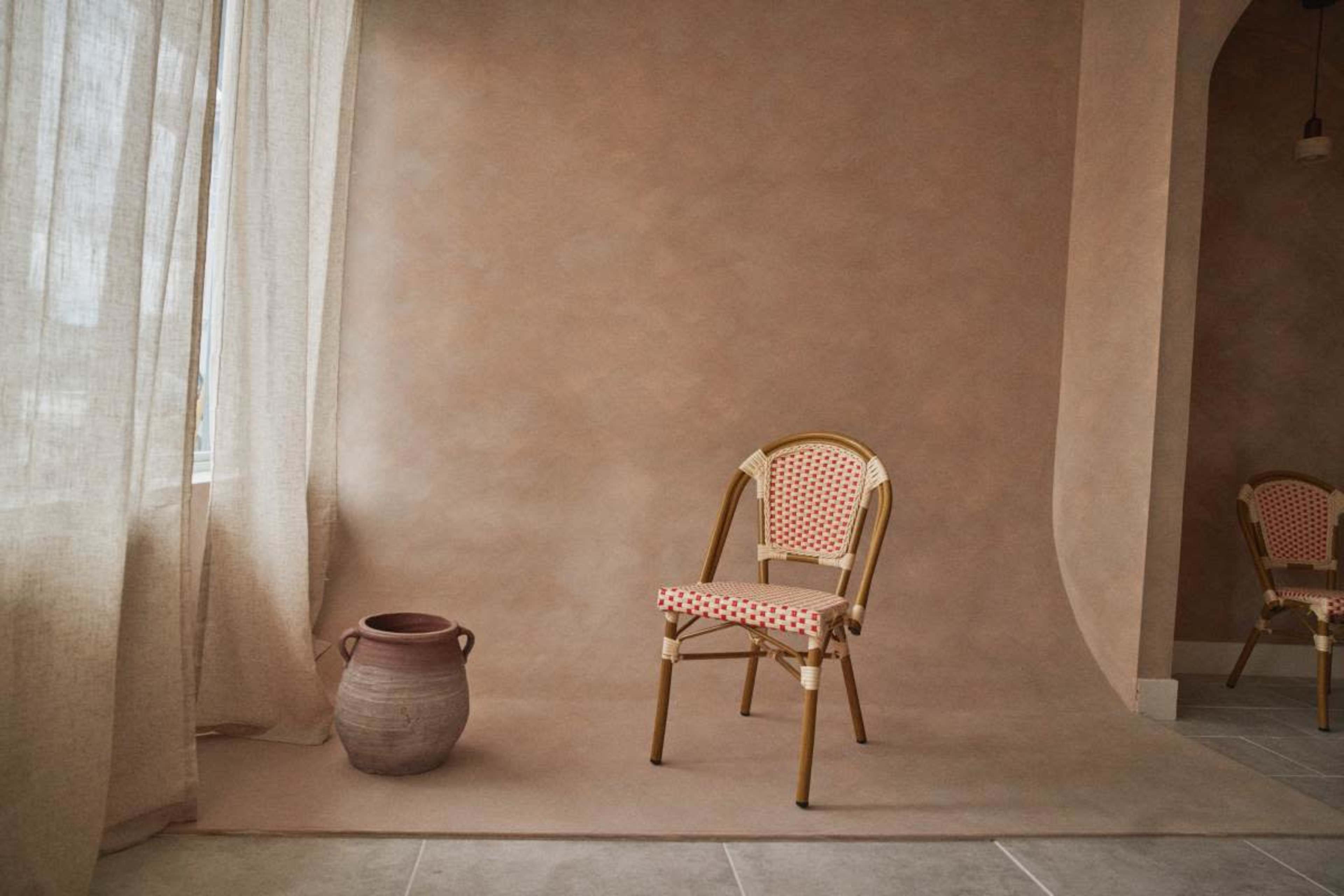 A handcrafted chair with a patterned seat is positioned beside a ceramic pot on a textured floor against a muted backdrop.