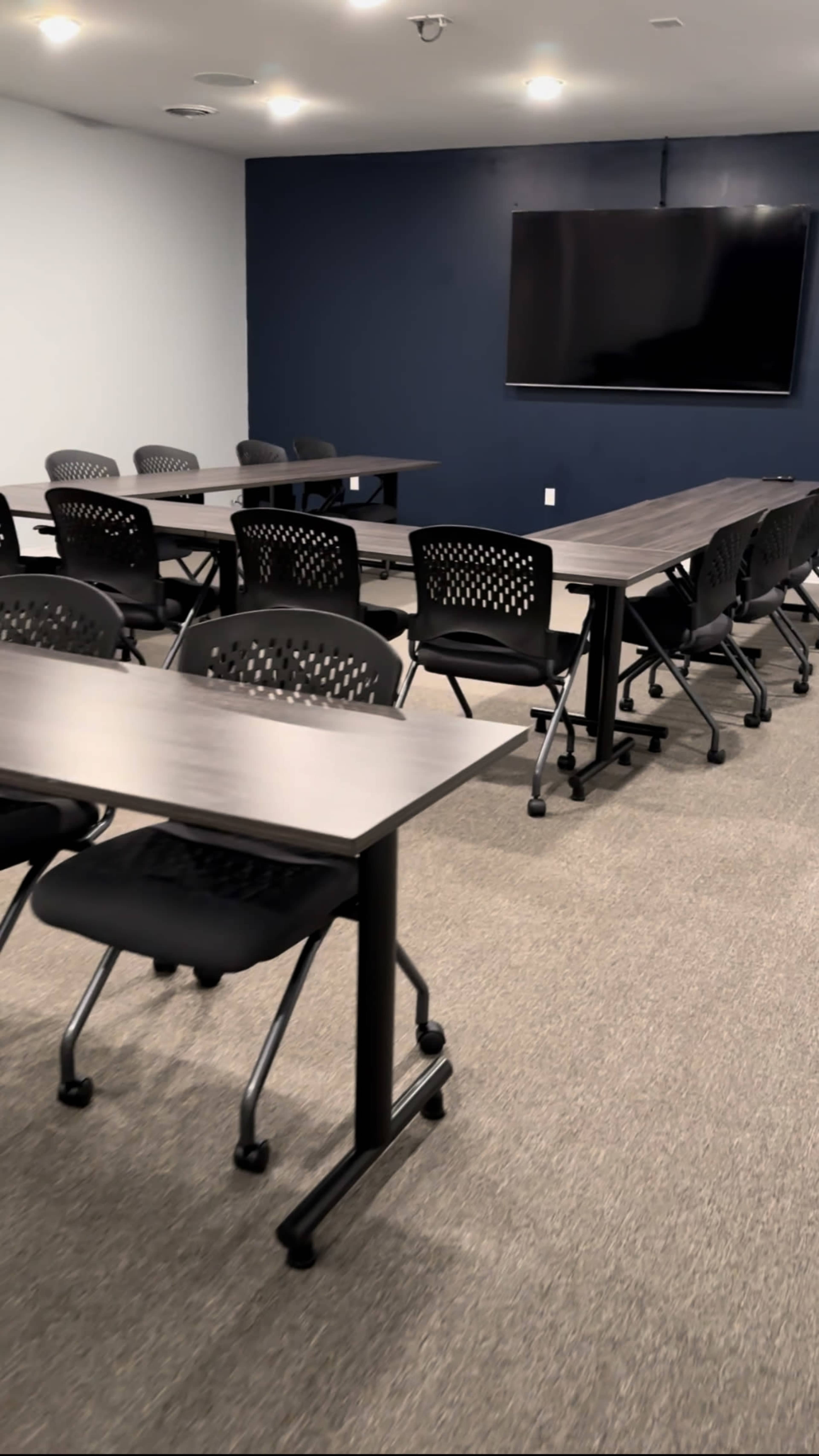A conference room features several arranged tables and rolling chairs with a television mounted on the wall.