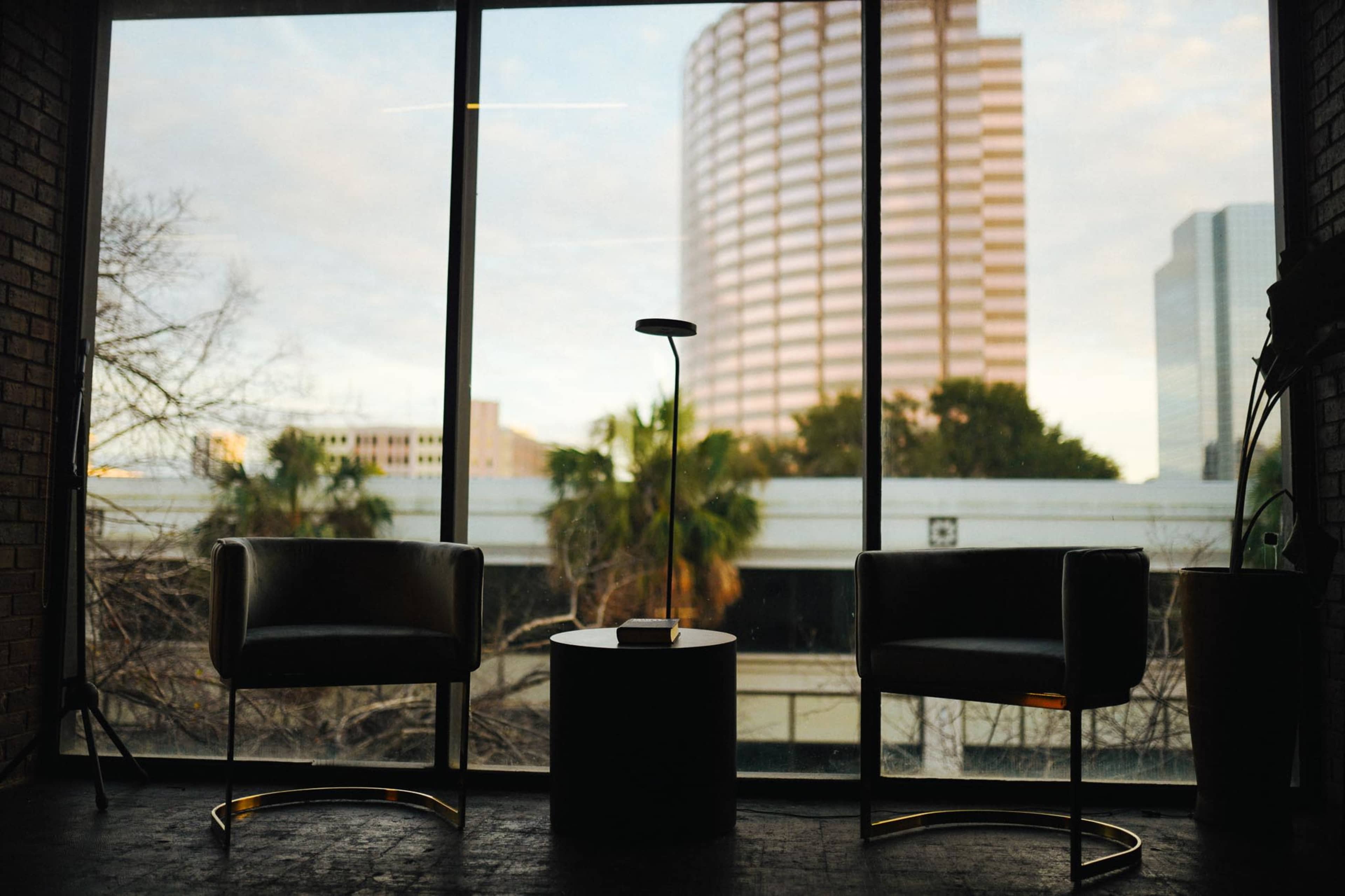 Two black chairs and a small table are positioned in front of a large window showing a city skyline with a pink-striped building and palm trees outside.