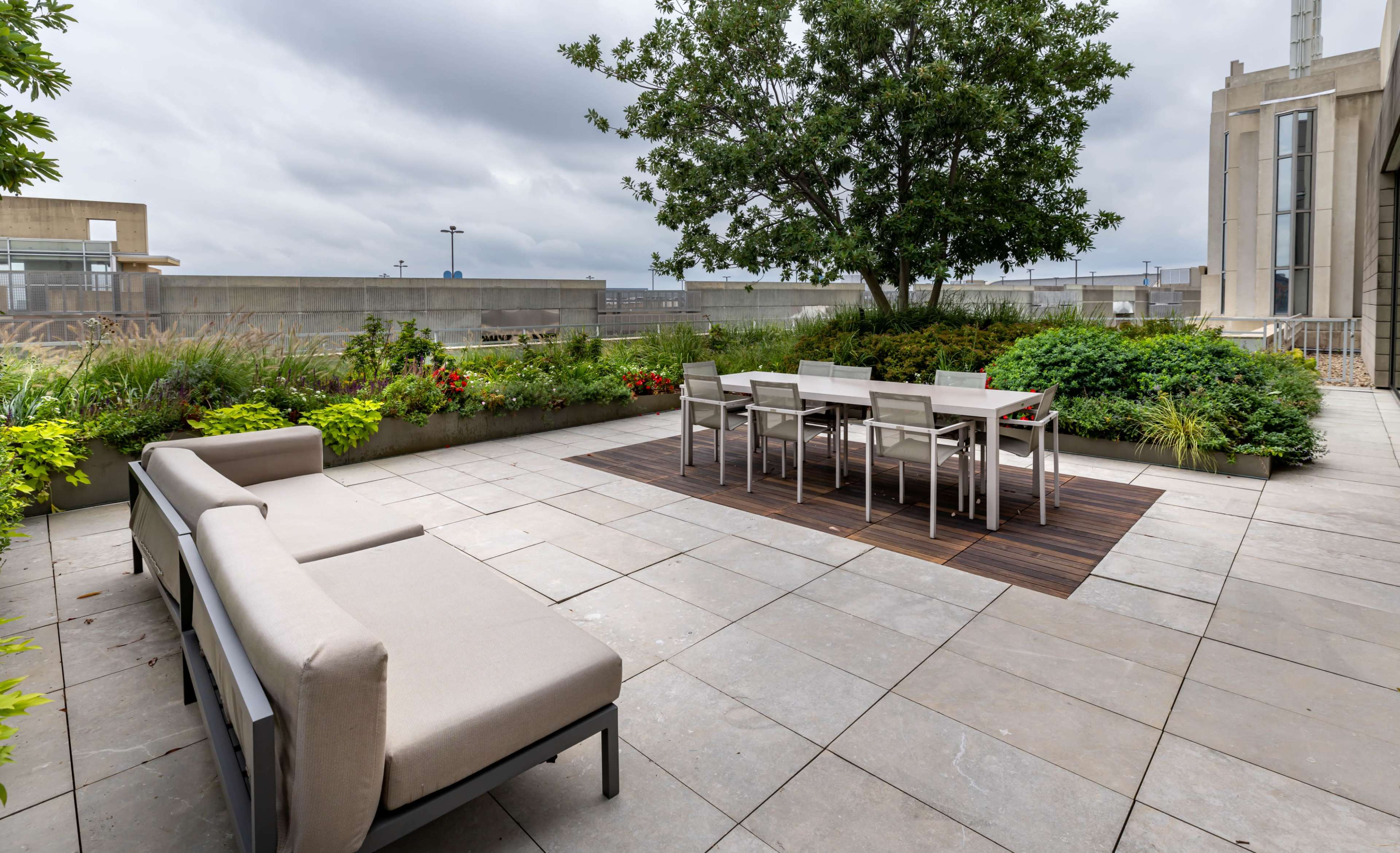 The image shows a spacious rooftop patio featuring a dining table with chairs, a seating area, and surrounding greenery under a cloudy sky.