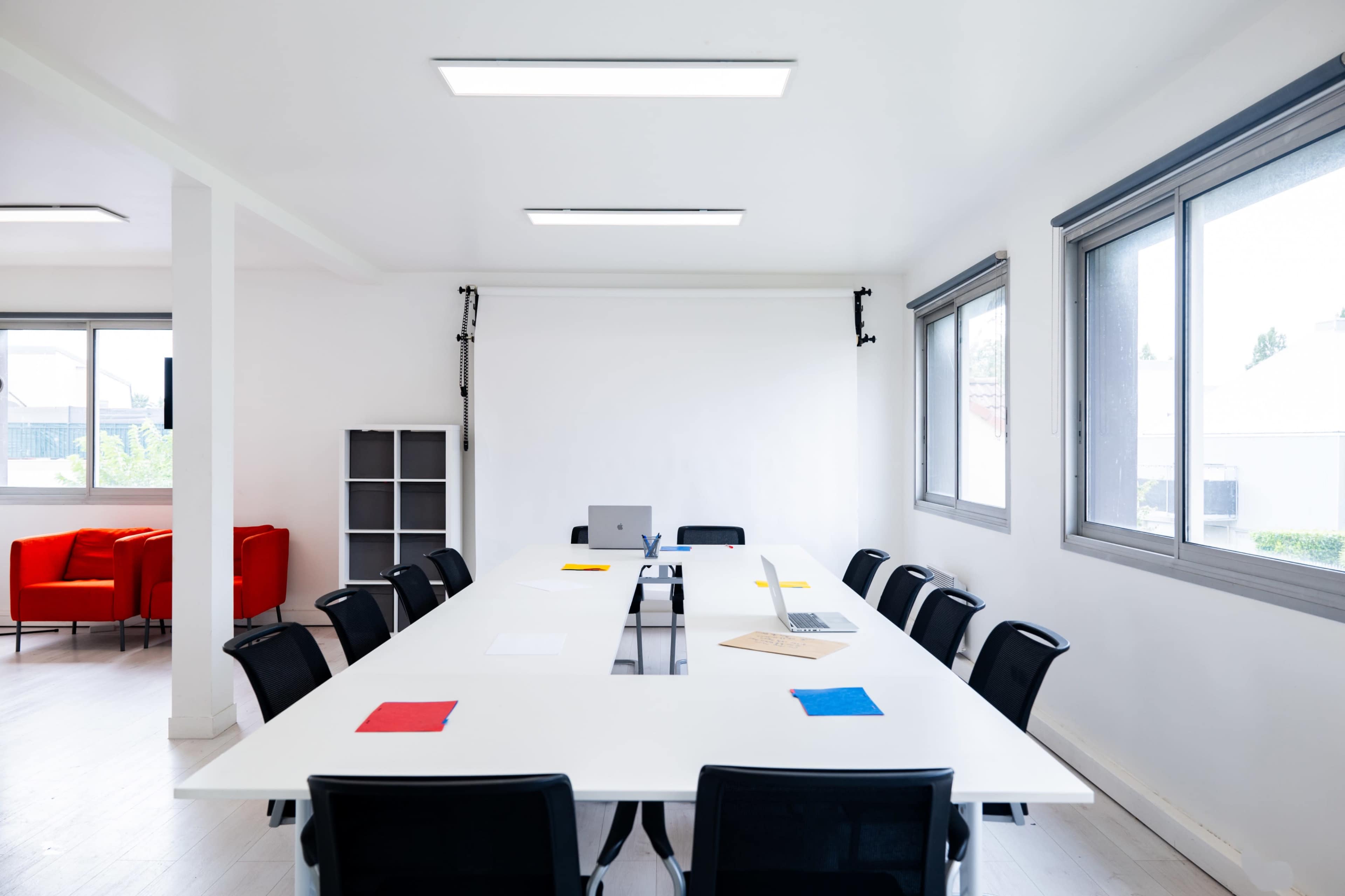 A large, modern meeting room features a long rectangular table with black chairs, a laptop, a monitor, and colored sheets of paper, all under bright overhead lighting and surrounded by windows.