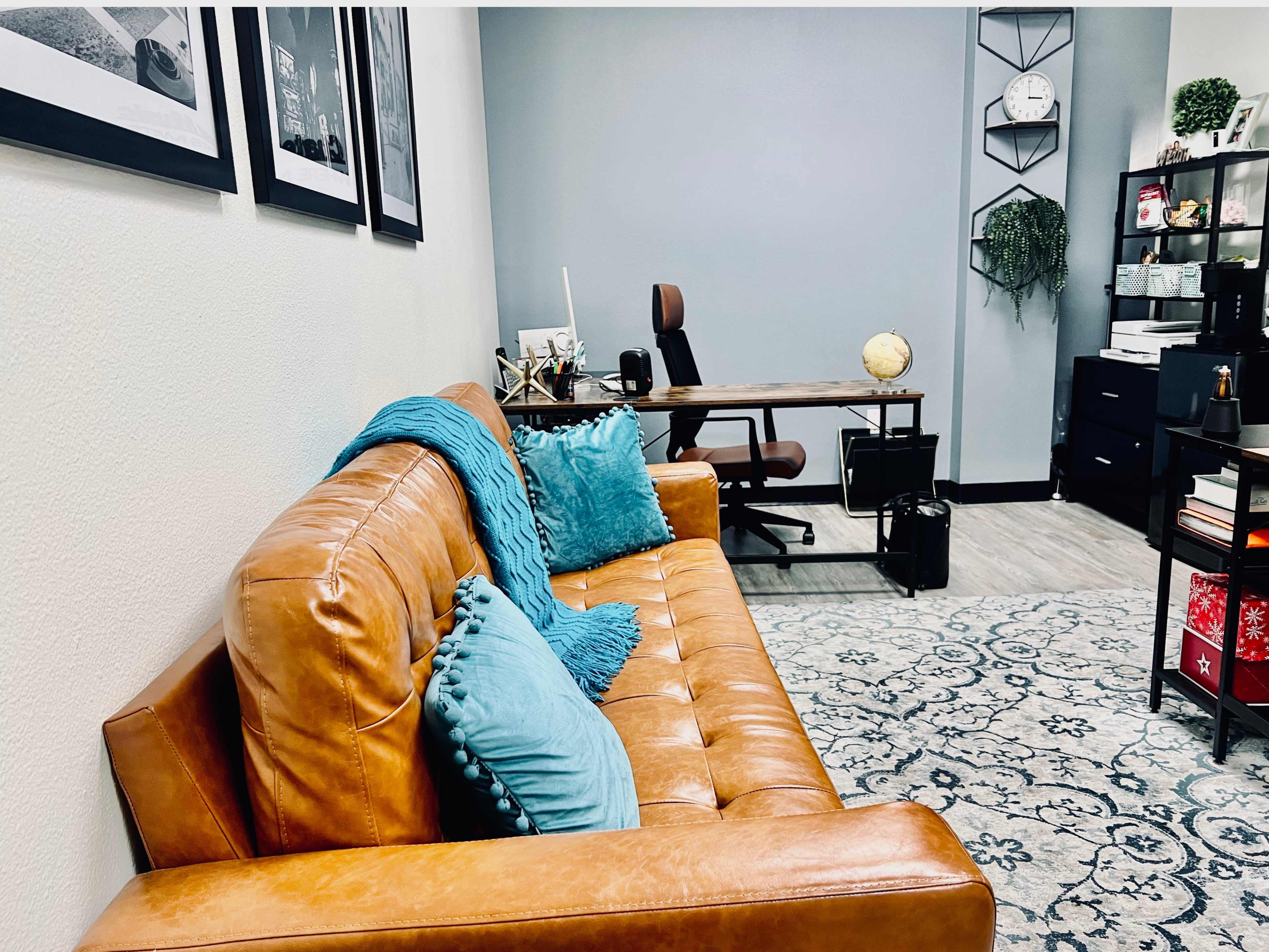 A modern office space featuring a brown leather sofa with turquoise pillows, a wooden desk with an office chair, and decorative elements on the walls and floor.