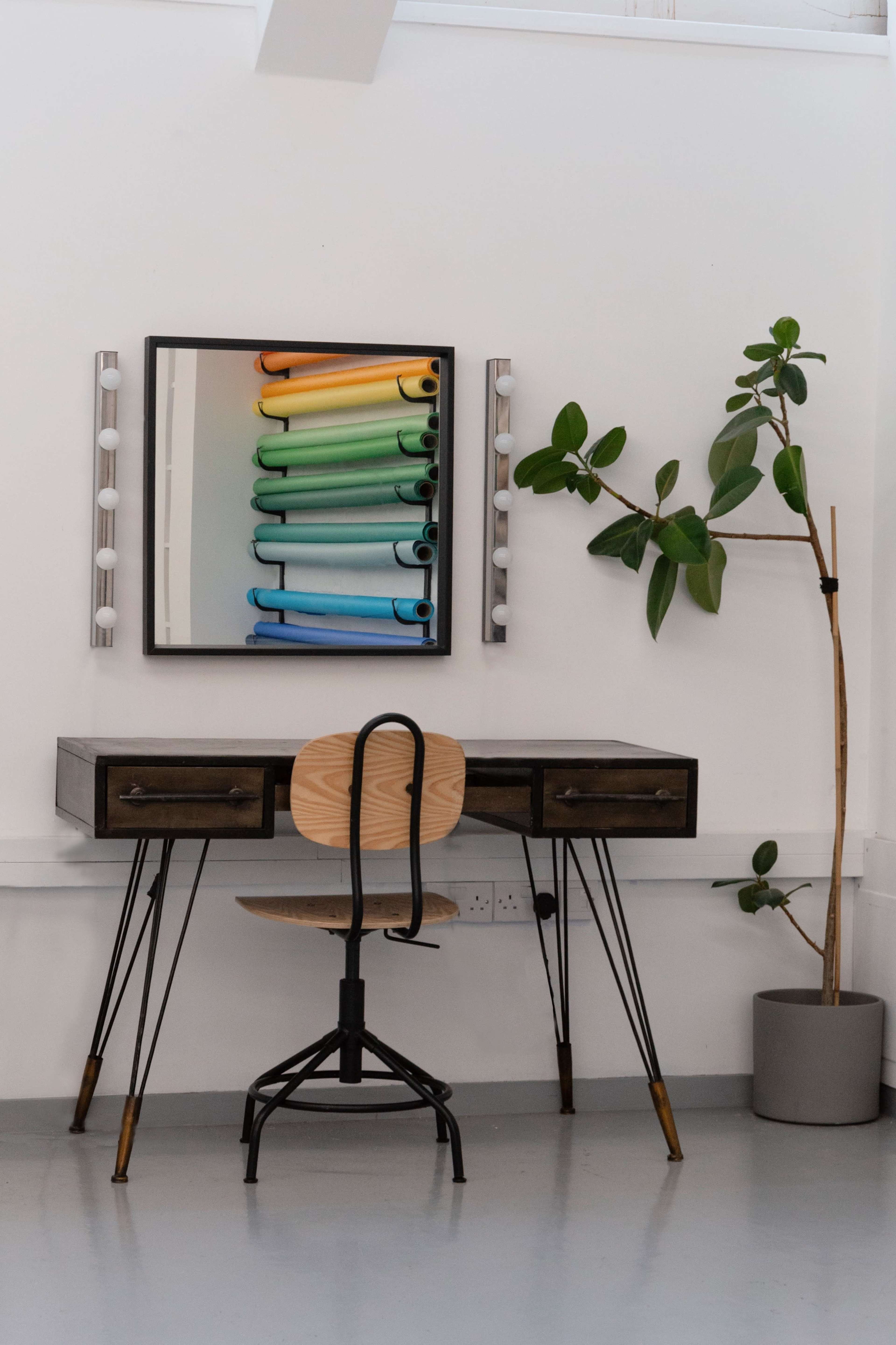 The image shows a minimalistic workspace featuring a wooden desk with drawers, a metal chair, a mirror reflecting colorful rolls of paper, and a potted plant in the corner.