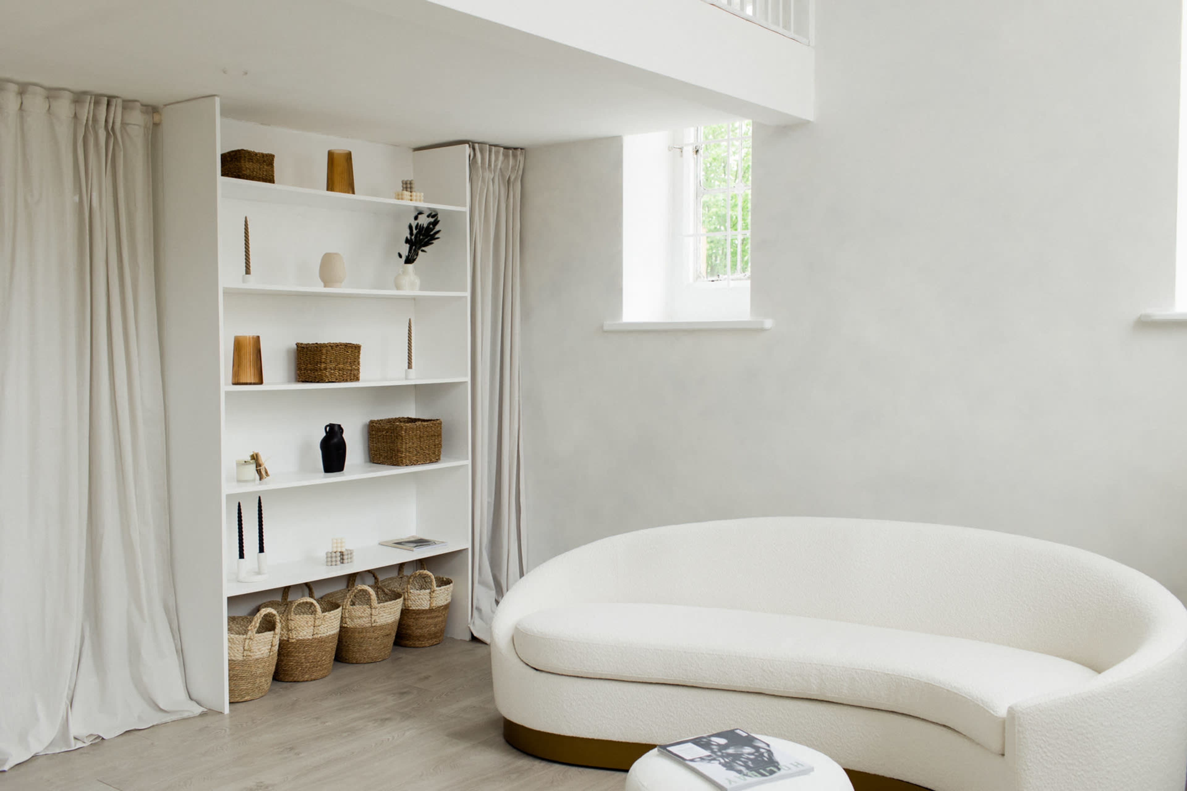 The image shows a modern living space with a curved white sofa, a sleek shelving unit filled with decorative items, and woven baskets on the floor, all set against light-colored walls.