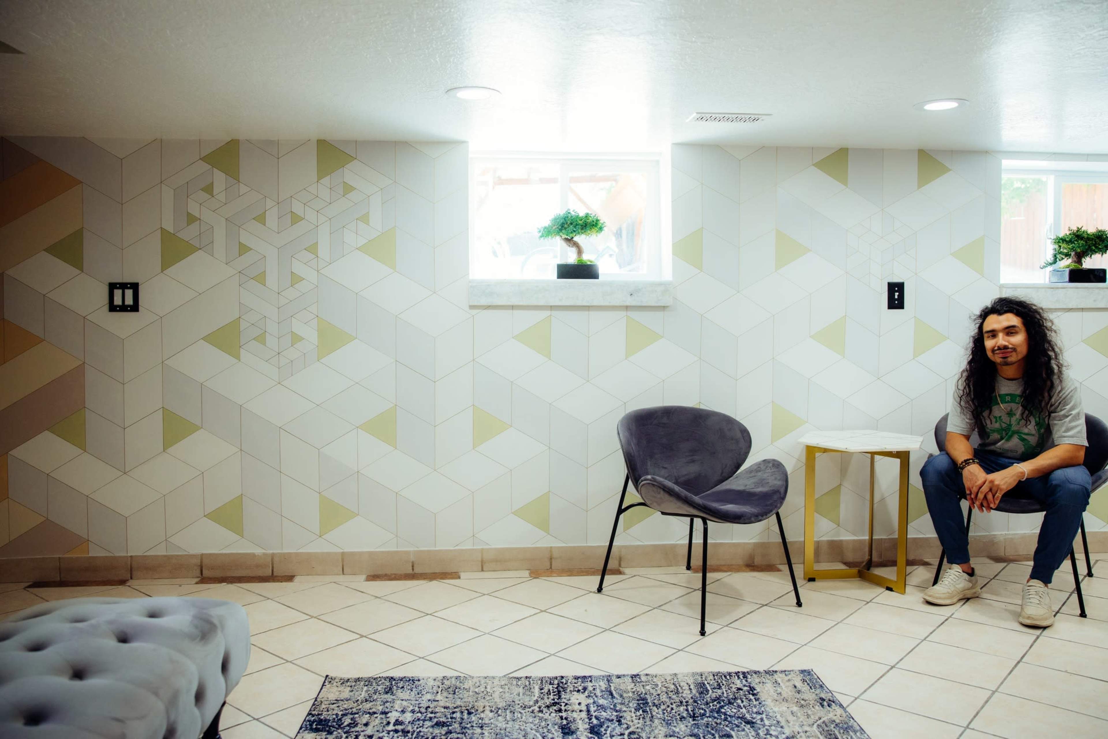 A man with long hair sits on a chair in a brightly lit room with geometric patterned walls and a tiled floor.