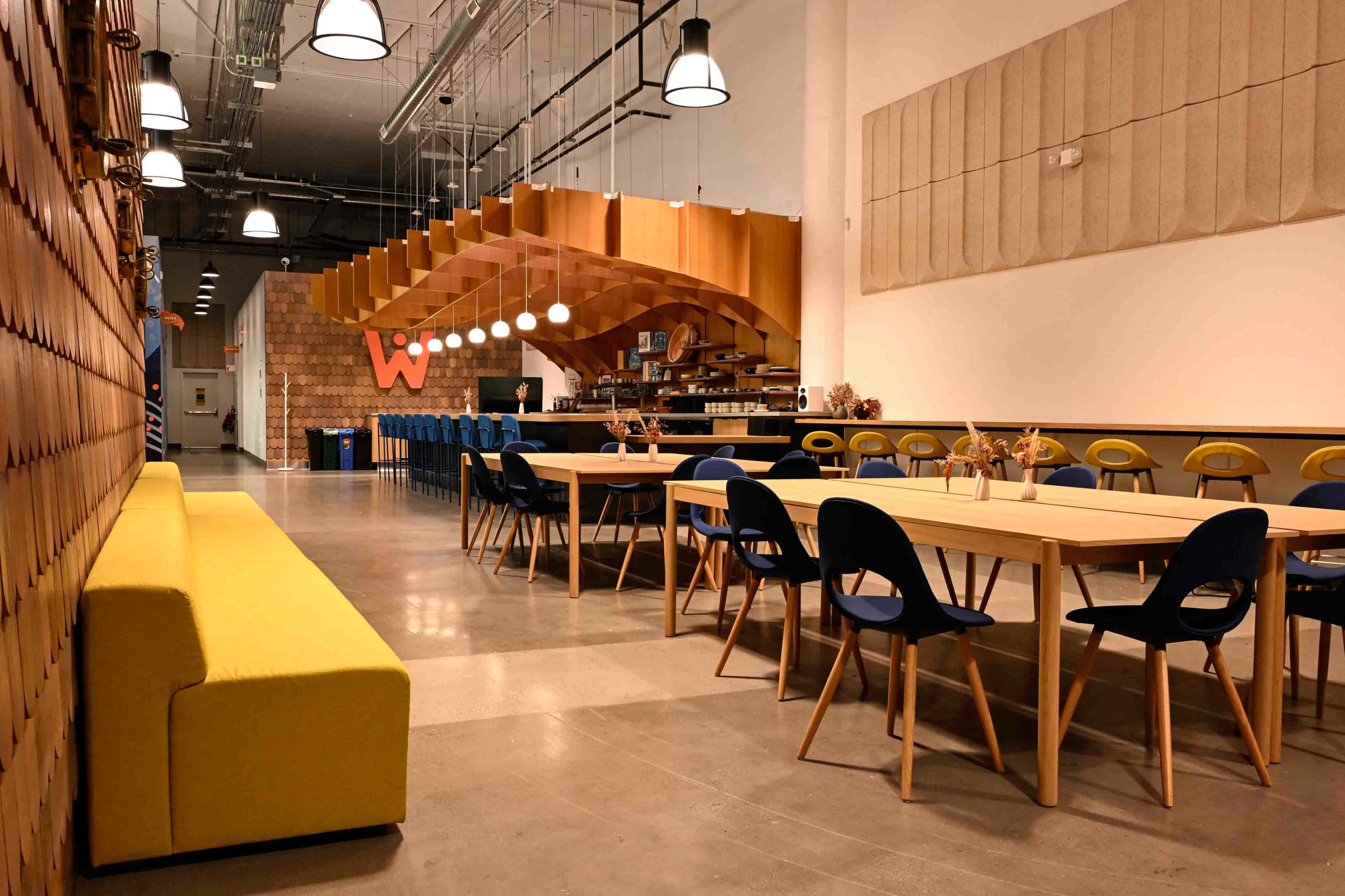 The image shows a modern, open dining area featuring a long wooden table with blue chairs, a yellow sofa along one wall, and wooden pendant lights above.