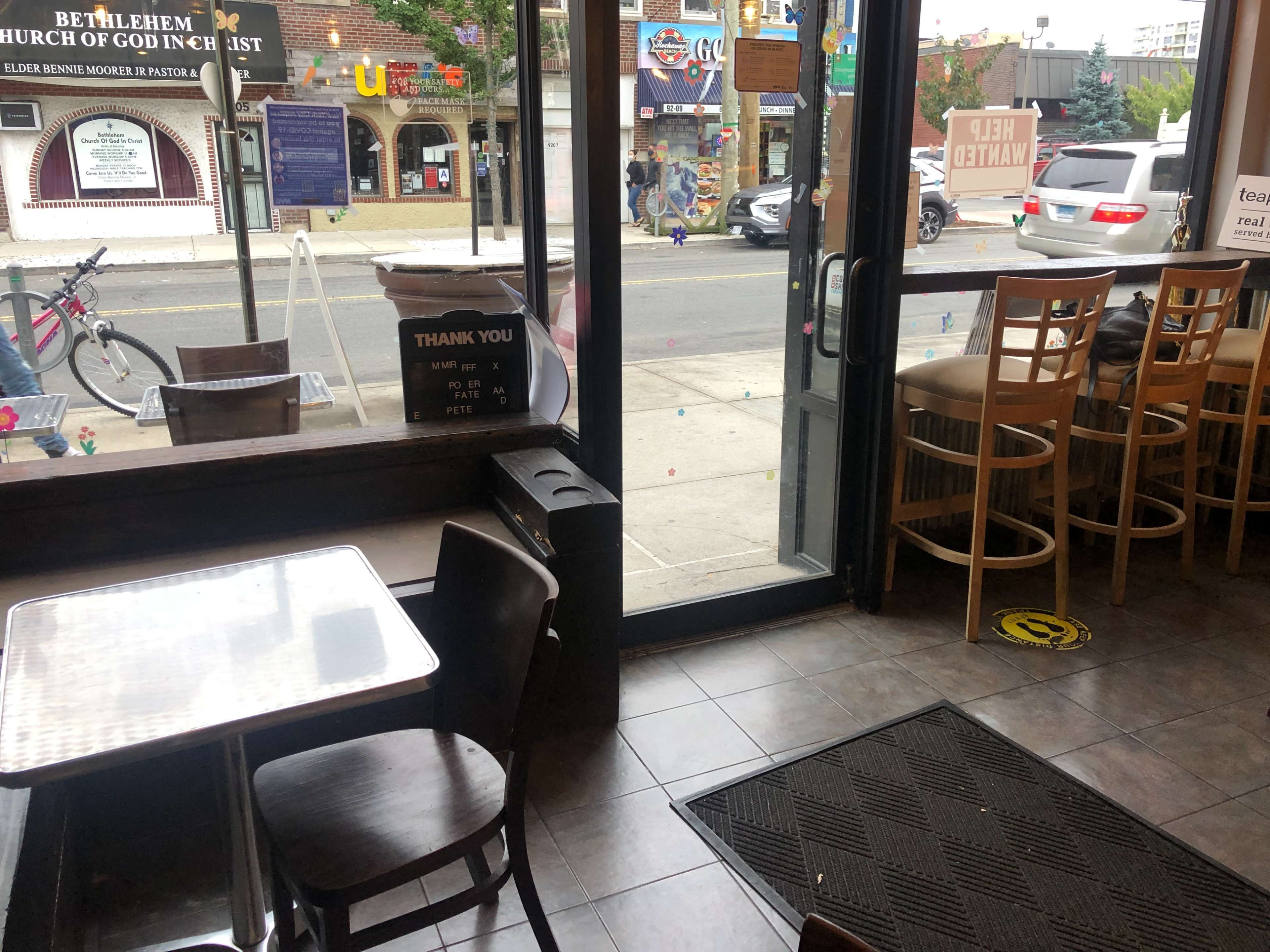 The inside of a café with empty seating, a door open to the street, and a visible 'Help Wanted' sign.