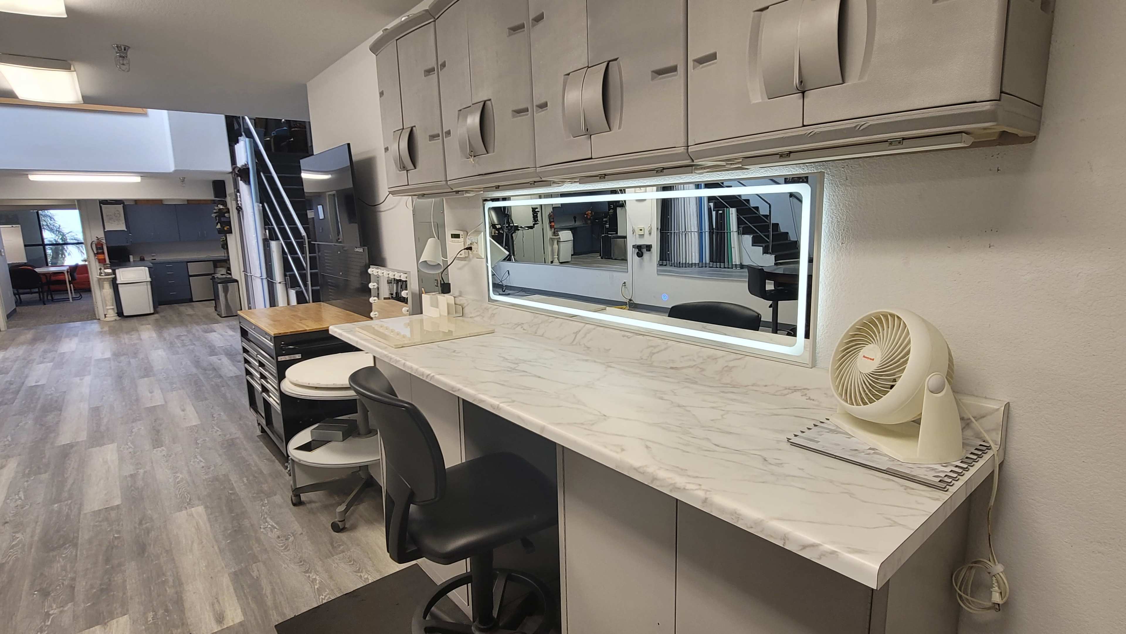A modern workspace with a long marble countertop, two black chairs, and overhead storage cabinets, illuminated by a backlit mirror.