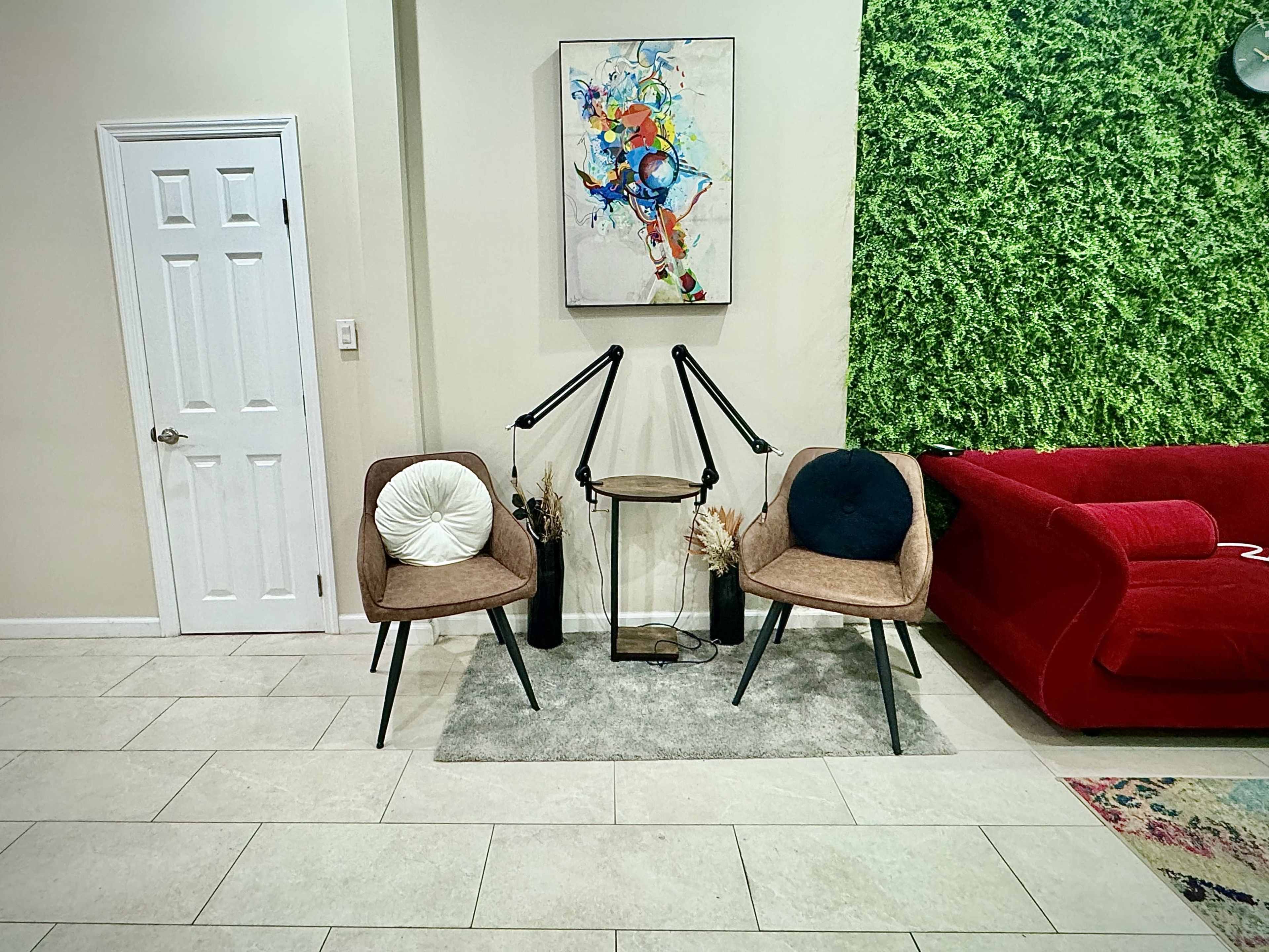 The image shows a living area with two modern chairs facing a small table, a vibrant piece of abstract art on the wall, and a green wall feature beside a red sofa.
