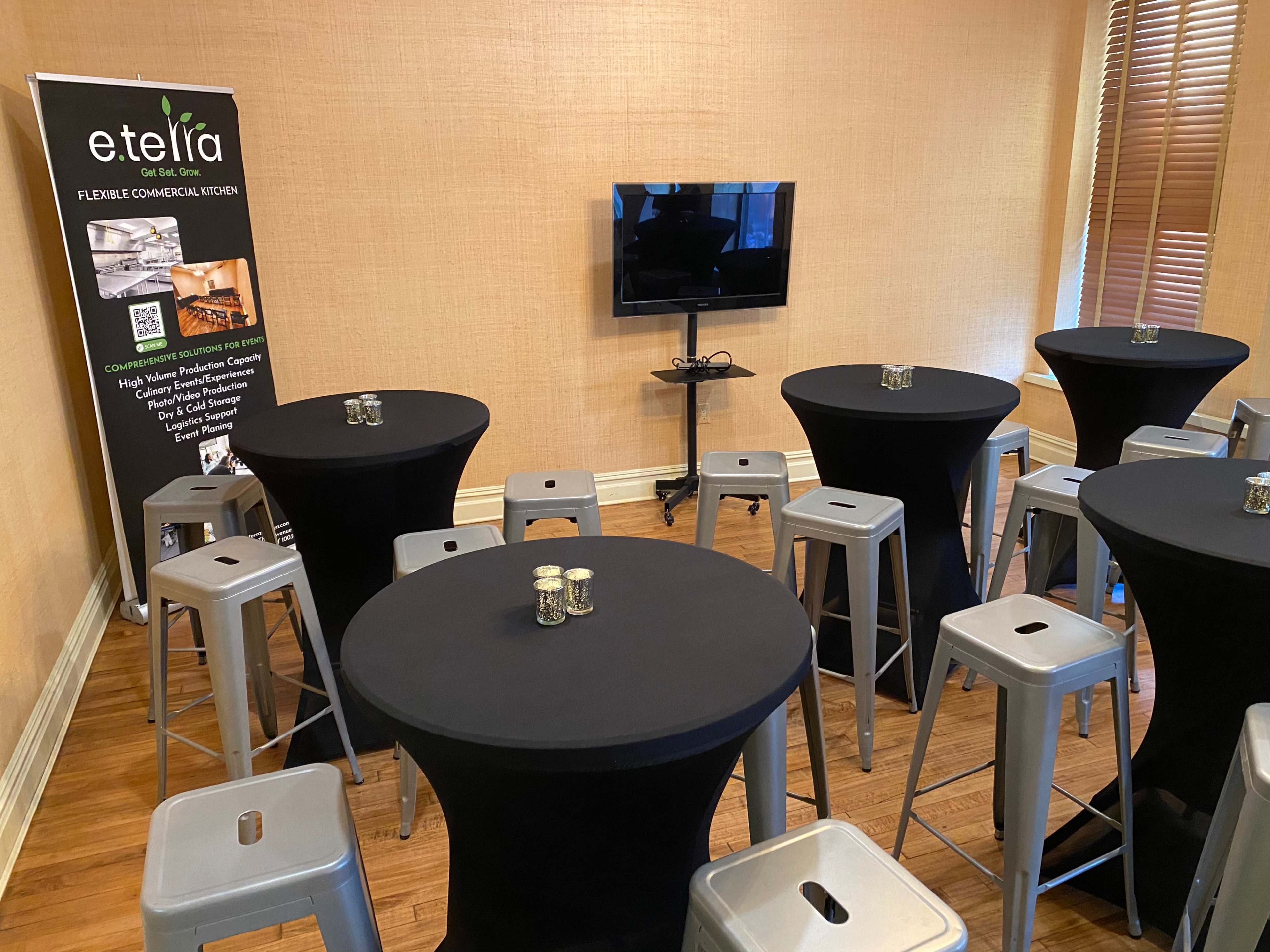 An interior space features several black tables surrounded by metal stools, with a television and a promotional banner in the background.