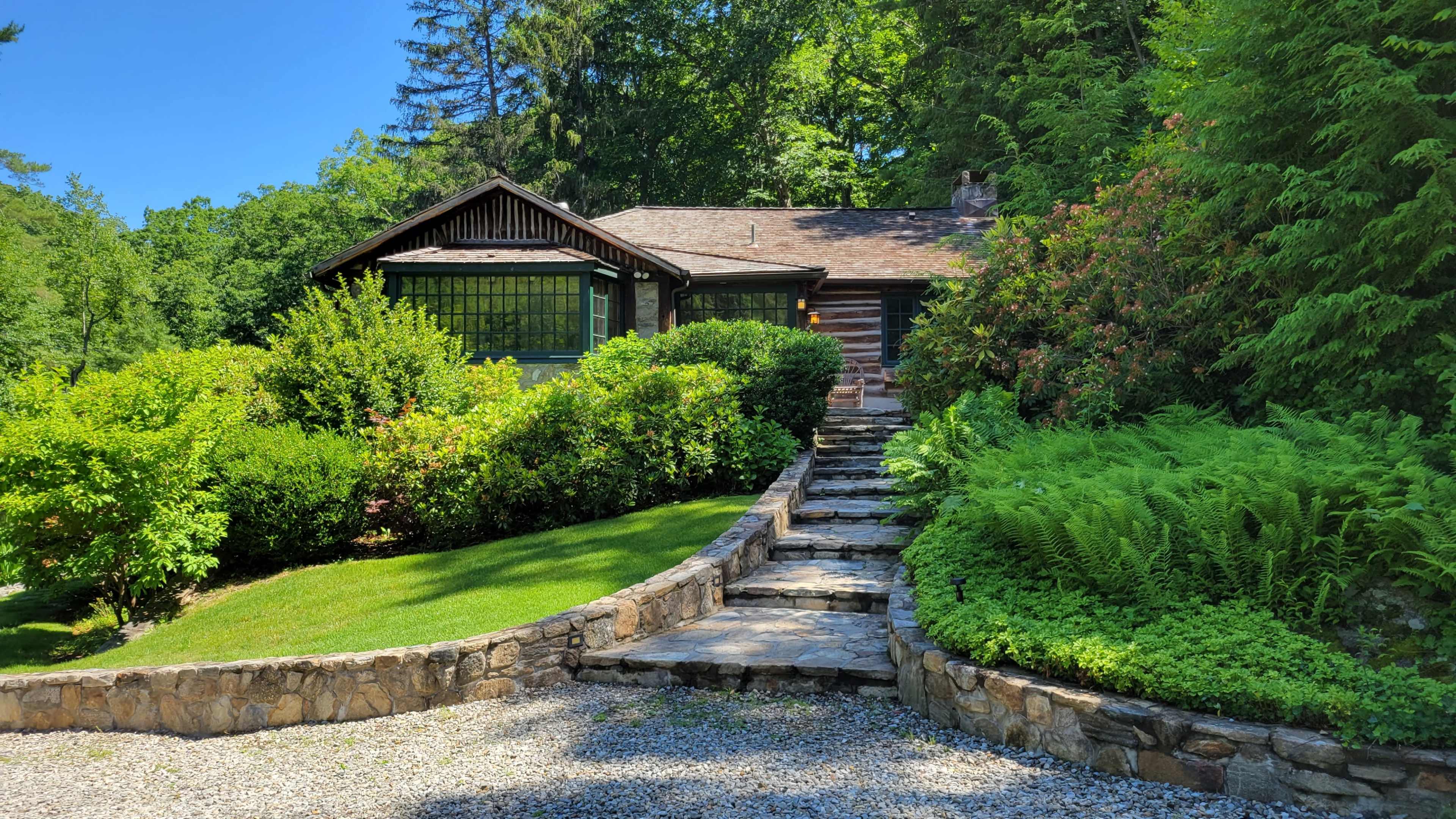 A stone pathway leads to a log cabin surrounded by lush greenery and flowering shrubs.