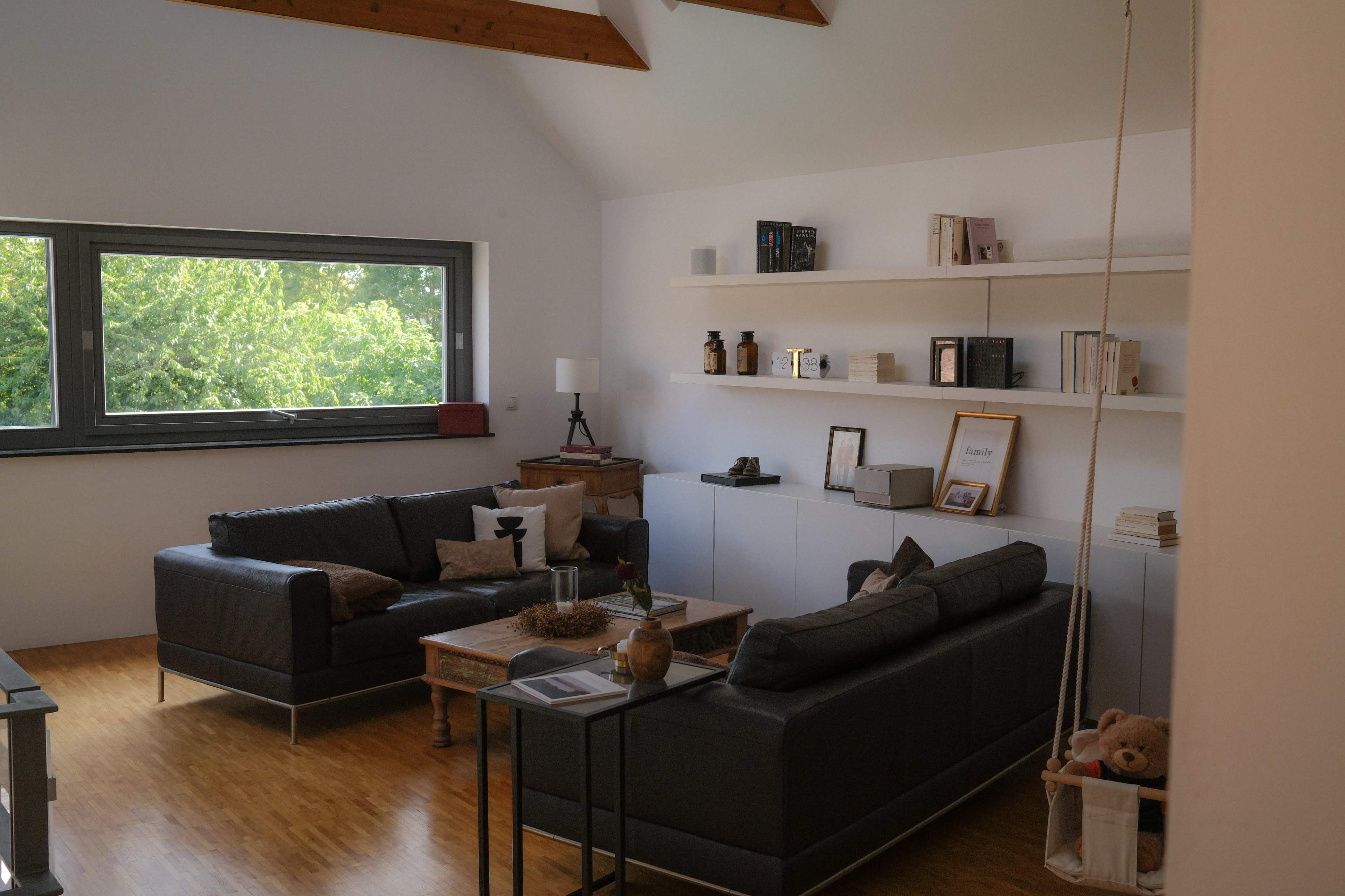 The image shows a bright, modern living room with two dark sofas, a coffee table, and shelves filled with books and decorative items against a backdrop of large windows.