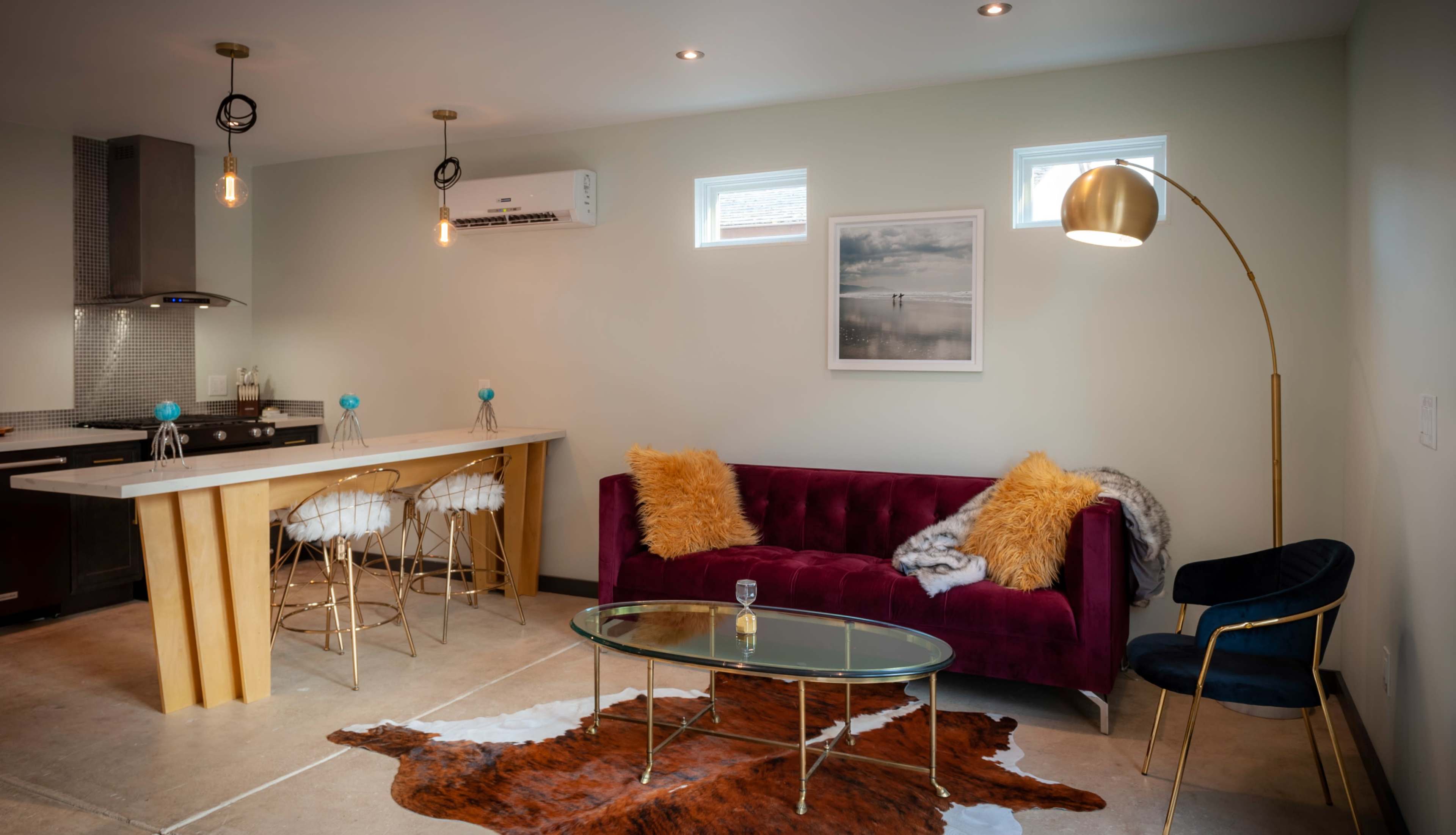 A modern living space featuring a purple velvet sofa, a glass coffee table on a cowhide rug, and a kitchen area in the background with a bar and stools.